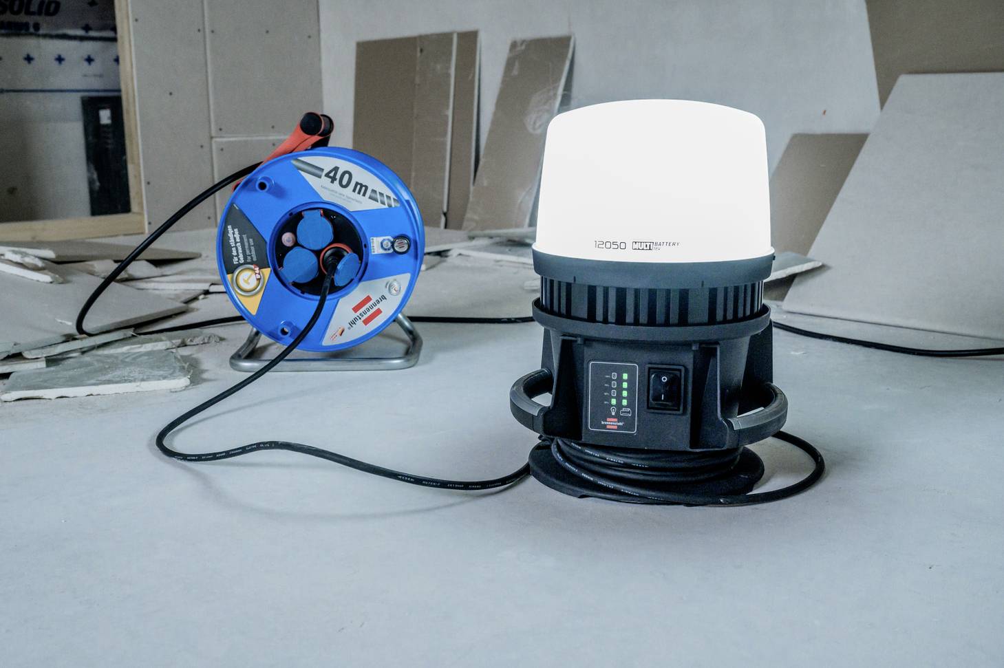 Eine Baustellenlampe leuchtet auf einem Boden, neben einer blauen Kabeltrommel mit der Aufschrift '40m'. Hintergrund aus Gipskartonplatten.