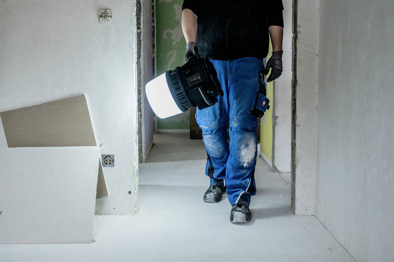 Eine Person in Baukleidung trägt eine helle Lampe durch einen unfertigen Raum. An den Wänden sind Steckdosen sichtbar.
