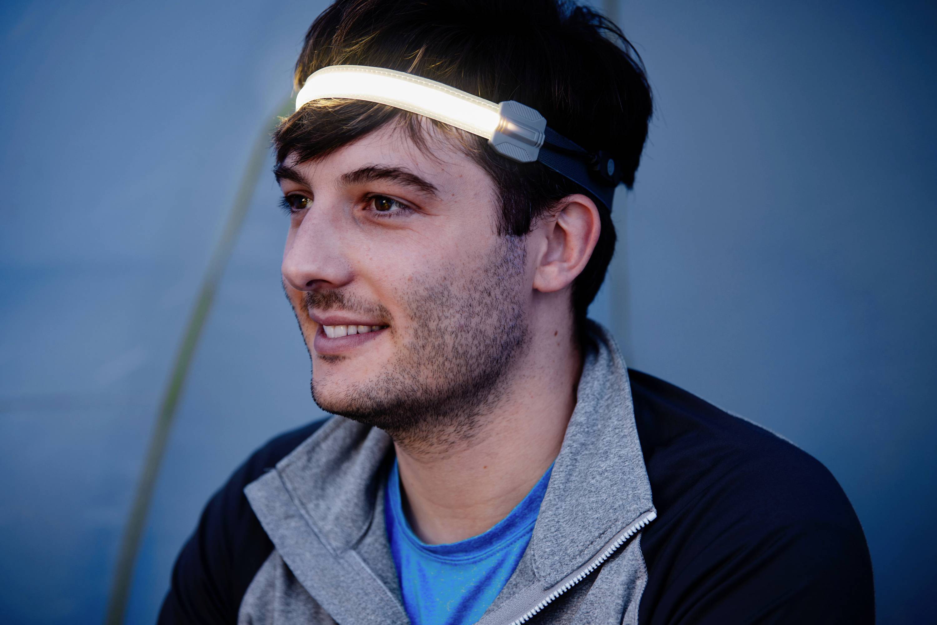 Ein lächelnder Mann trägt ein Stirnband mit einer leuchtenden LED. Hintergrund ist verschwommen und blau.