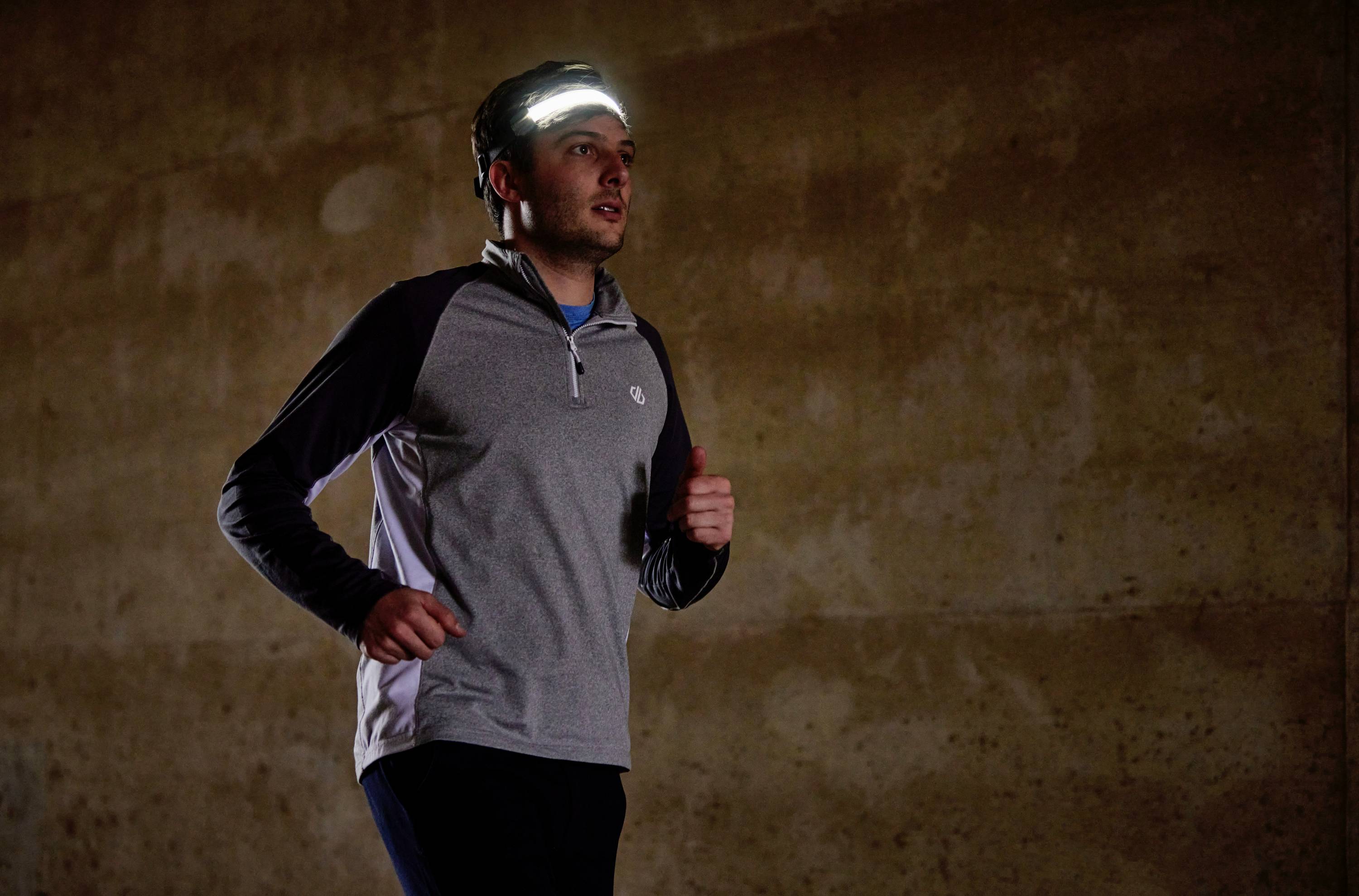 Ein Mann joggt in einem dunklen Raum, trägt ein langärmeliges Shirt und eine Stirnlampe, die seine Umgebung beleuchtet.