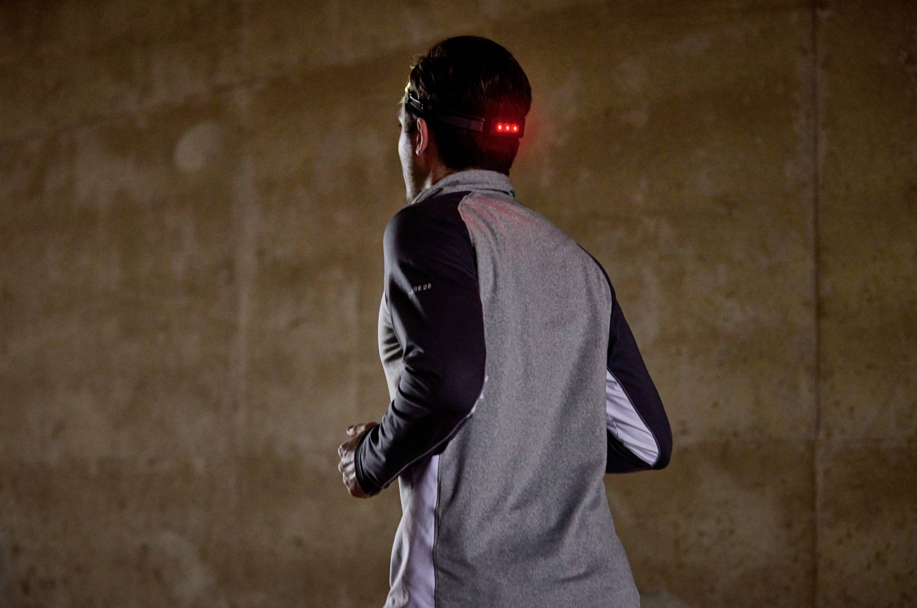 Eine Person joggt in einem dunklen Raum, trägt ein graues Oberteil mit reflektierenden Elementen und eine leuchtende LED-Lampe auf dem Kopf.