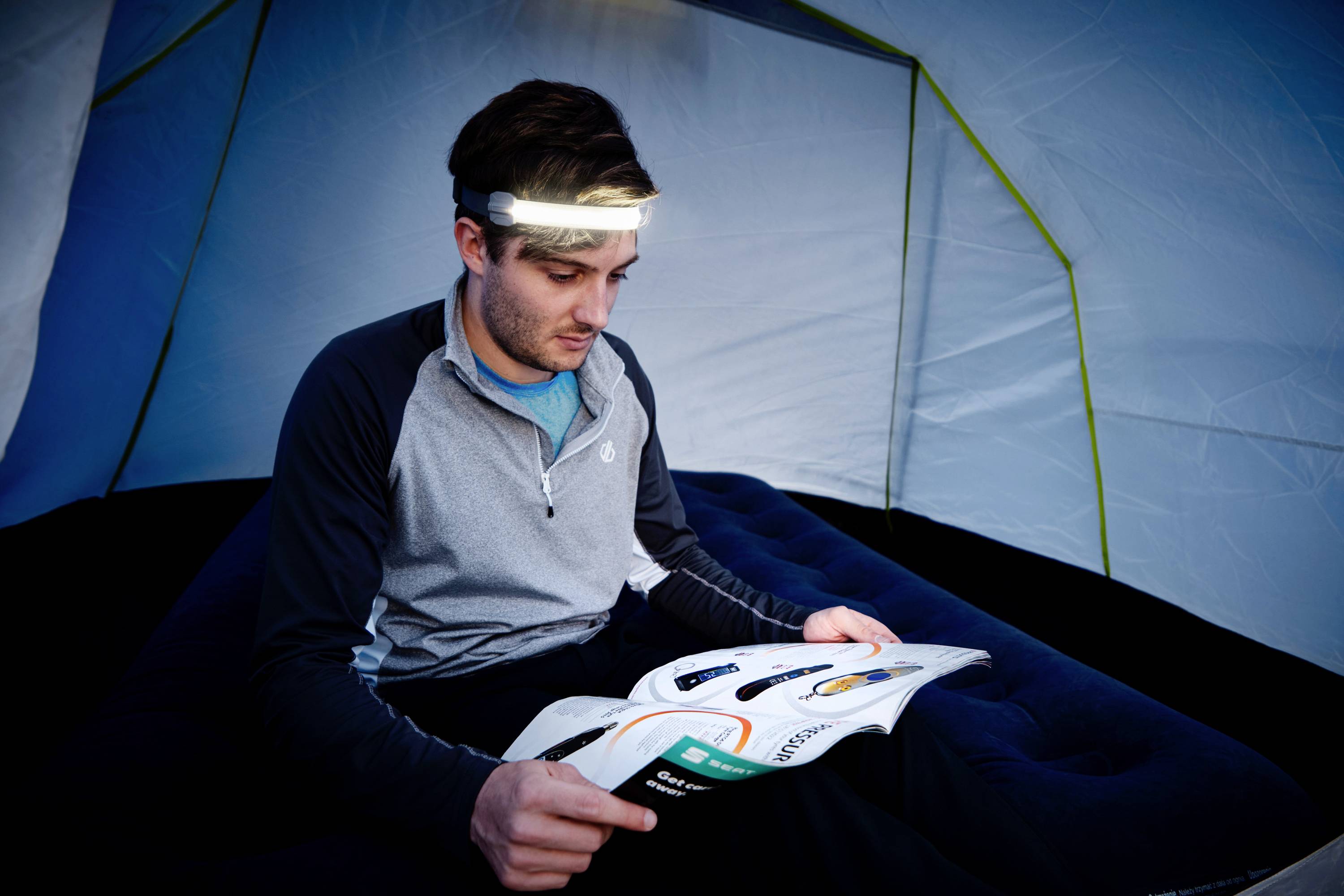 Eine Person sitzt in einem Zelt auf einer Luftmatratze und liest ein Buch mit einer Stirnlampe auf dem Kopf.