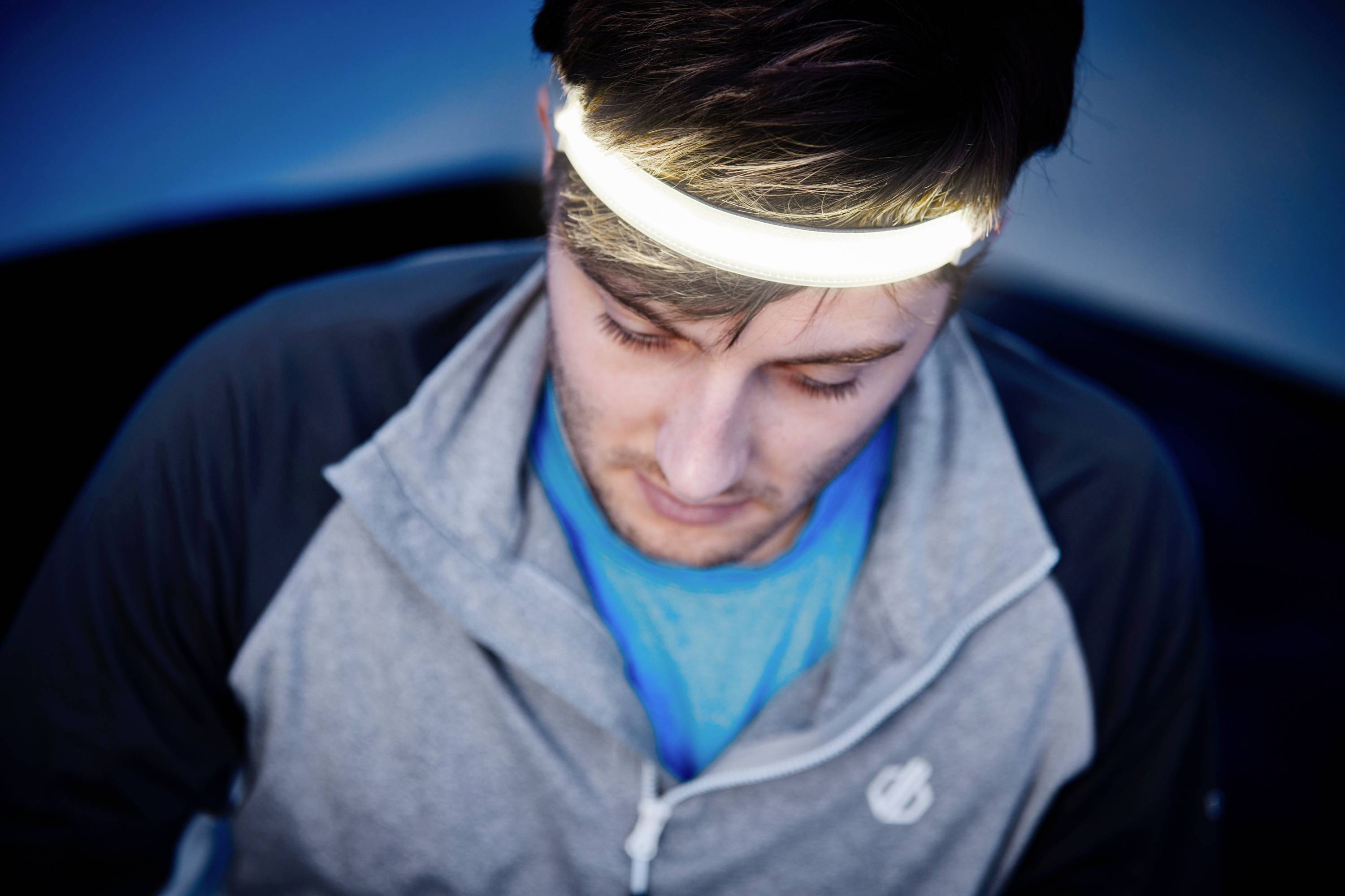 Ein Mann mit einer grauen Jacke trägt ein leuchtendes Stirnband. Sein Blick ist nach unten gerichtet. Hintergrund dunkelblau.