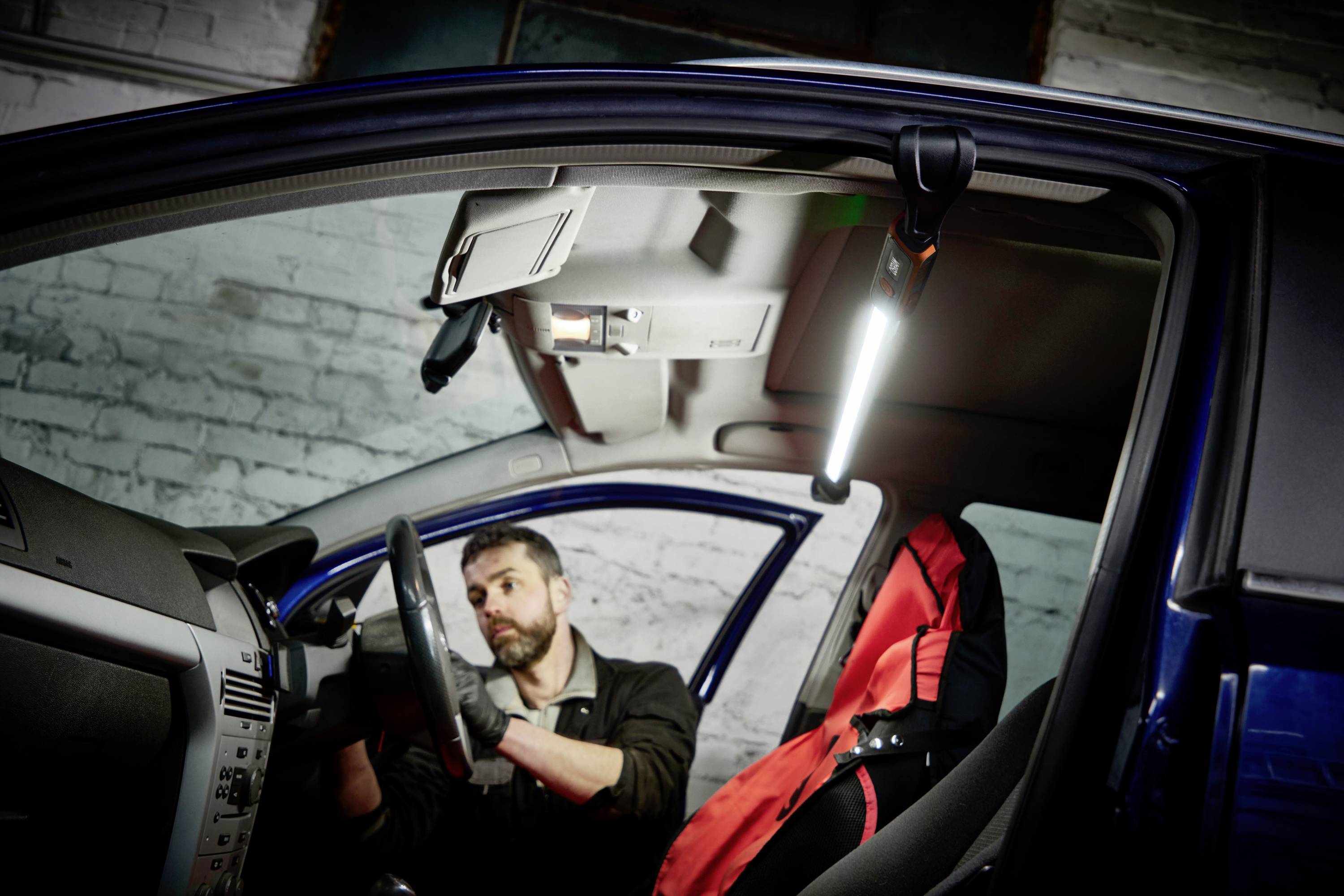 Ein Mechaniker arbeitet im Inneren eines Autos. Er trägt Handschuhe und nutzt eine helle Lampe zur Beleuchtung des Innenraums.