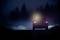 Ein Auto fährt nachts auf einer dunklen Straße, umgeben von Nebel. Das Fernlicht leuchtet den Weg aus, während Bäume die Straße säumen.