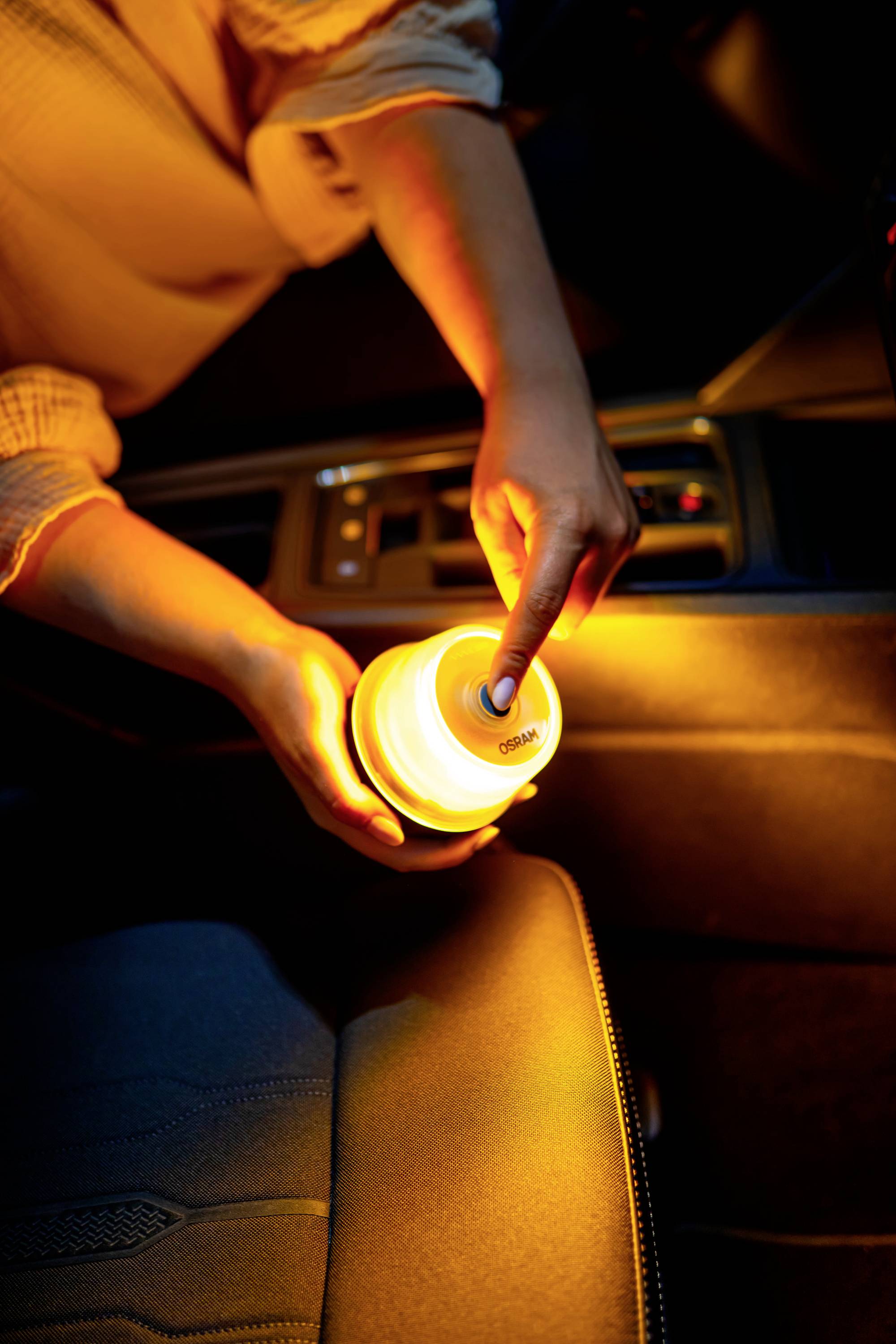 Eine Person schaltet in einem Auto eine tragbare, leuchtende Lampe ein. Die Szene ist in warmem, gelbem Licht beleuchtet.