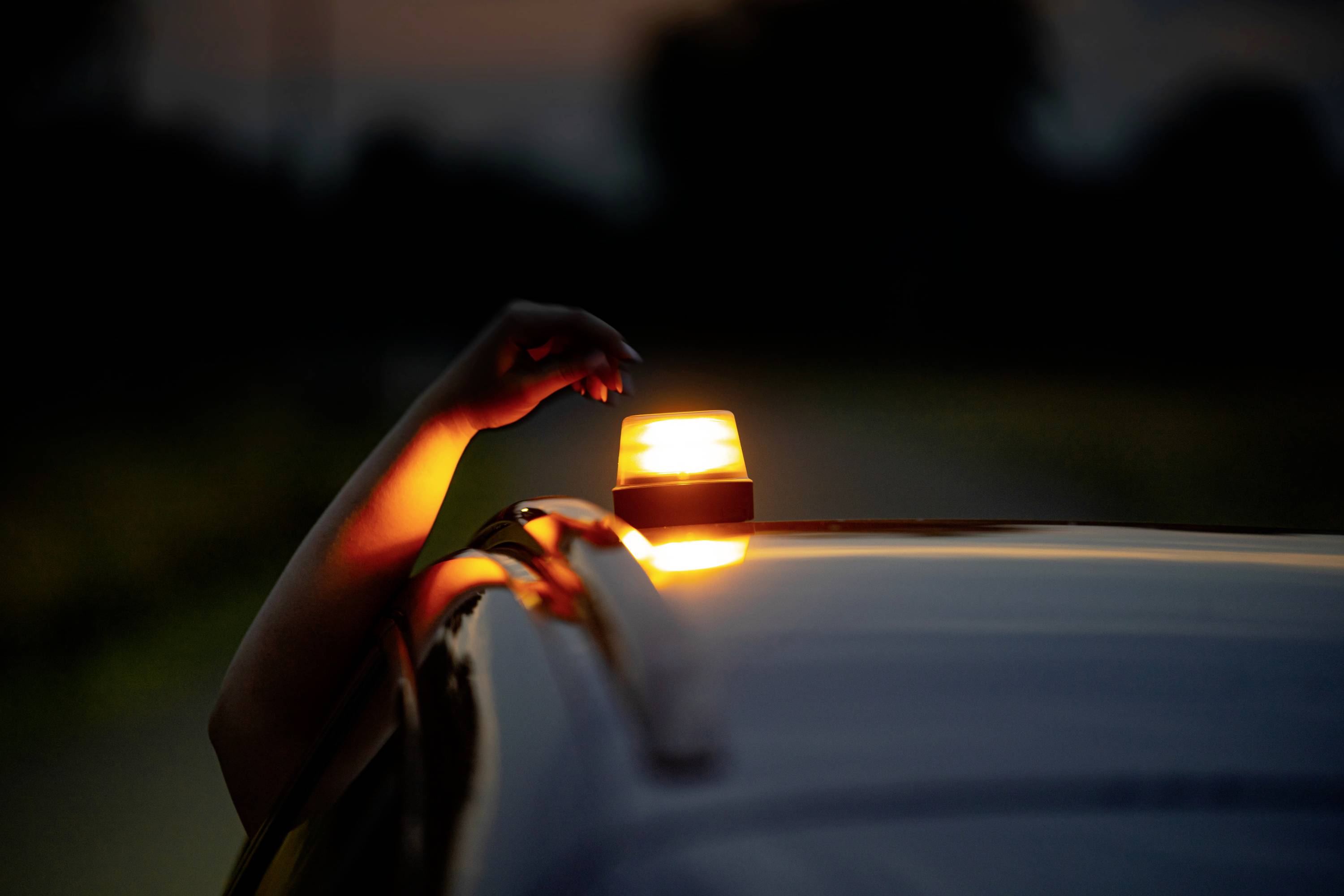 Eine Hand reicht aus einem Auto heraus und bewegt ein orangefarbenes Warnlicht auf dem Dach. Die Umgebung ist dunkel.