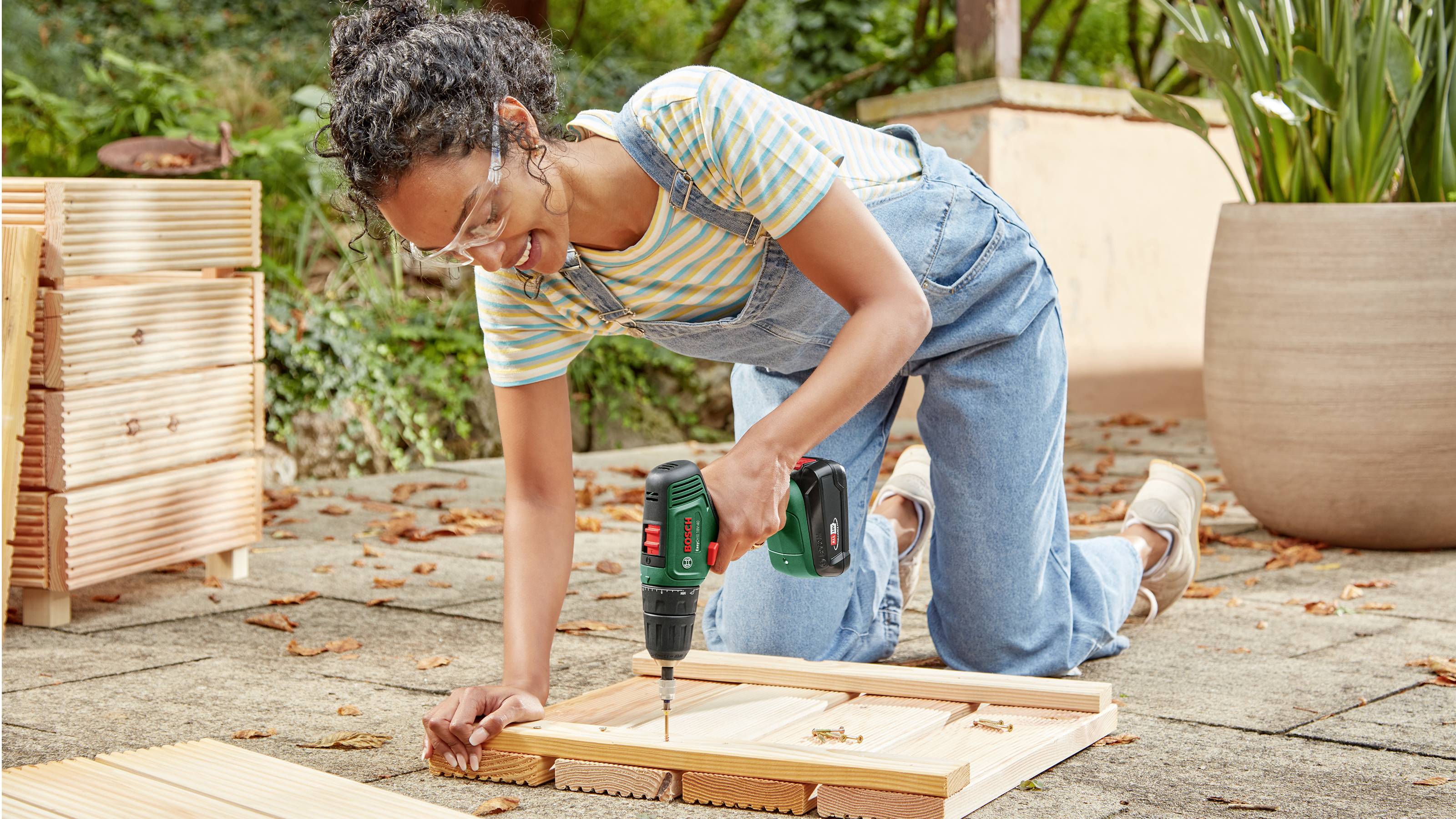 Eine Person kniet im Freien, trägt Schutzbrille und arbeitet mit einem Akkubohrer an Holzplatten, um möglicherweise einen Rahmen oder eine Struktur zu bauen.