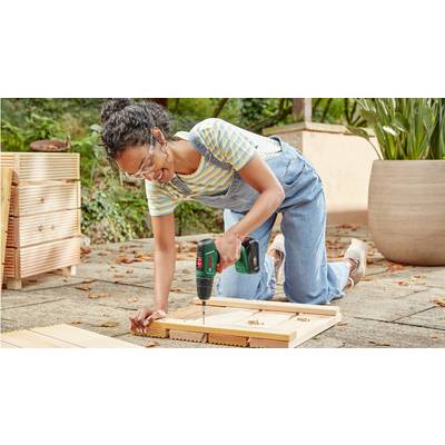 Eine Person kniet im Freien, trägt Schutzbrille und arbeitet mit einem Akkubohrer an Holzplatten, um möglicherweise einen Rahmen oder eine Struktur zu bauen.