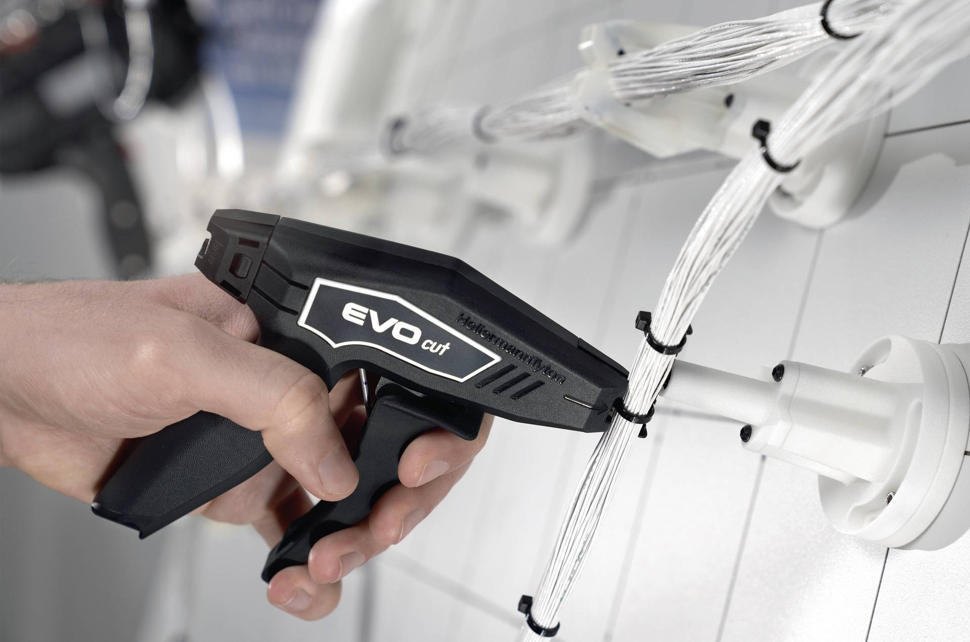 Eine Hand hält ein Werkzeug mit der Aufschrift 'Evo cut', das Kunststoffkabelbinder an einer weißen Kabelverbindung schneidet.