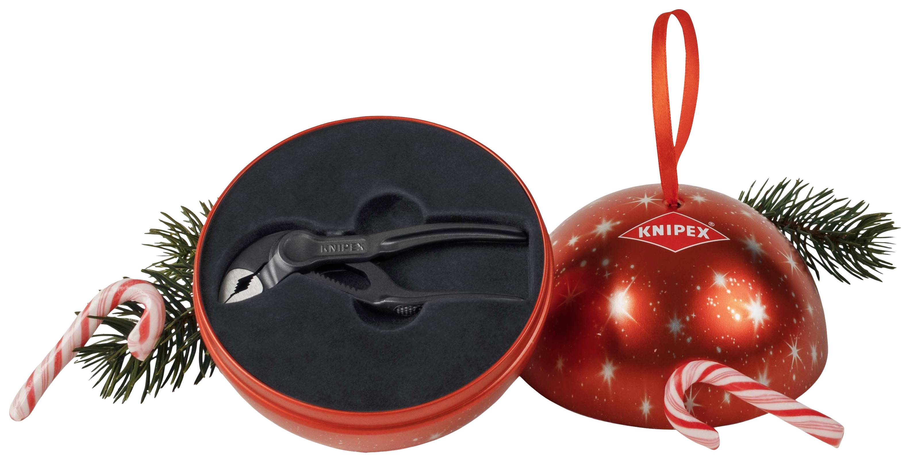 Ein rotes, kugelförmiges Weihnachtsgeschenk-Set von Knipex, geöffnet, zeigt einen schwarzen Zangenschlüssel. Dekoriert mit Tannenzweigen und Zuckerstangen.