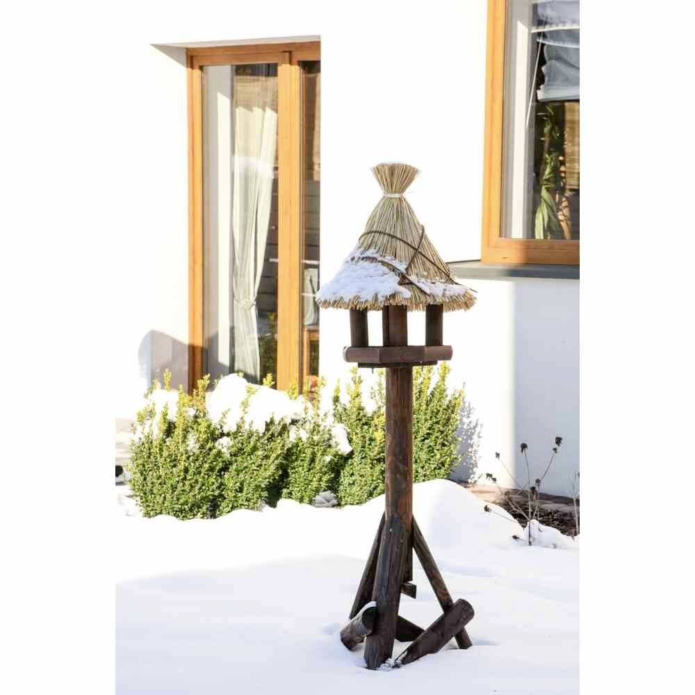 Ein hölzernes Vogelhäuschen mit Strohdach steht im verschneiten Garten vor einem Haus mit Holzfenstern. Sträucher im Hintergrund.