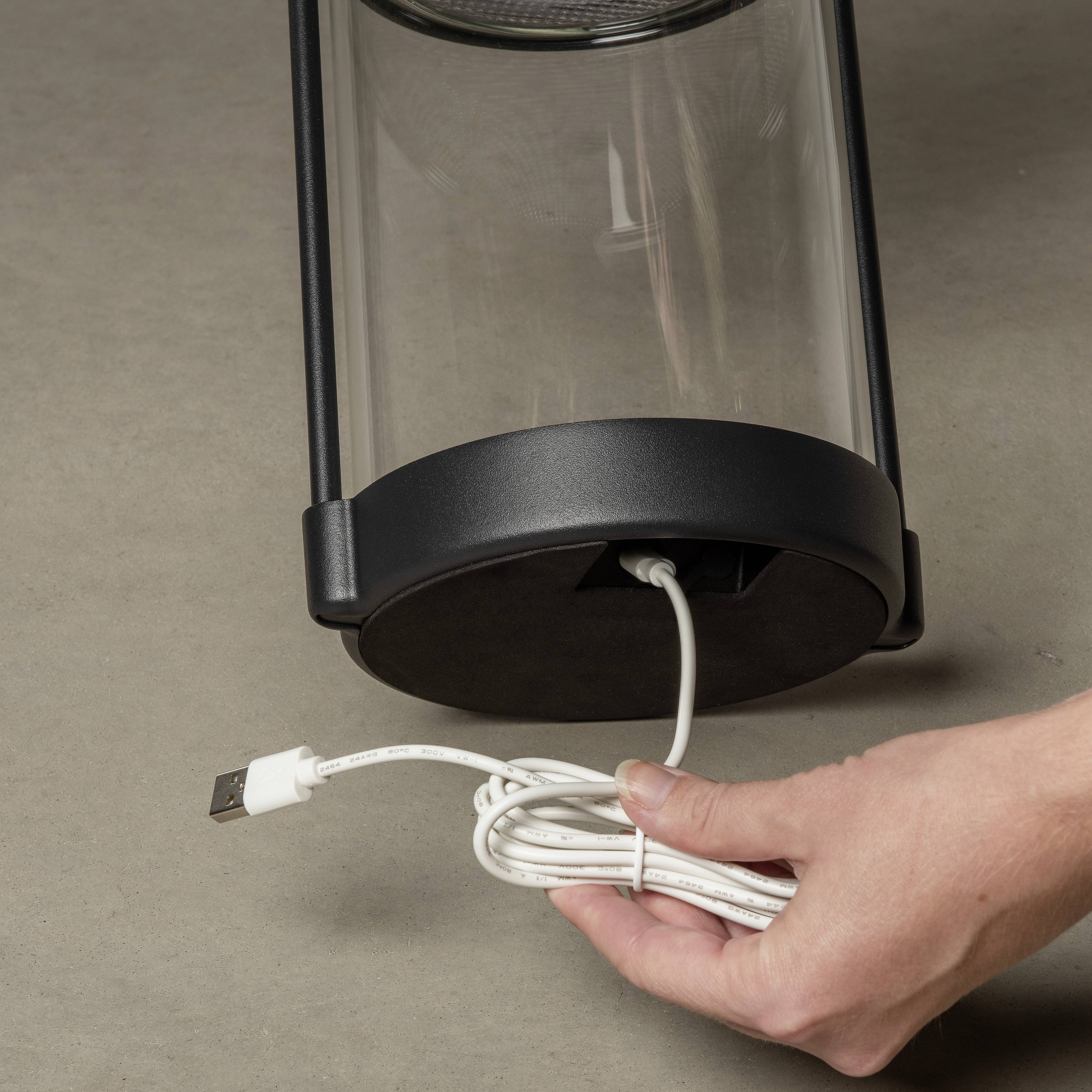 Eine Hand hält ein Kabel, das mit der Unterseite eines zylinderförmigen Geräts verbunden ist. Das Gerät steht auf einem grauen Untergrund.