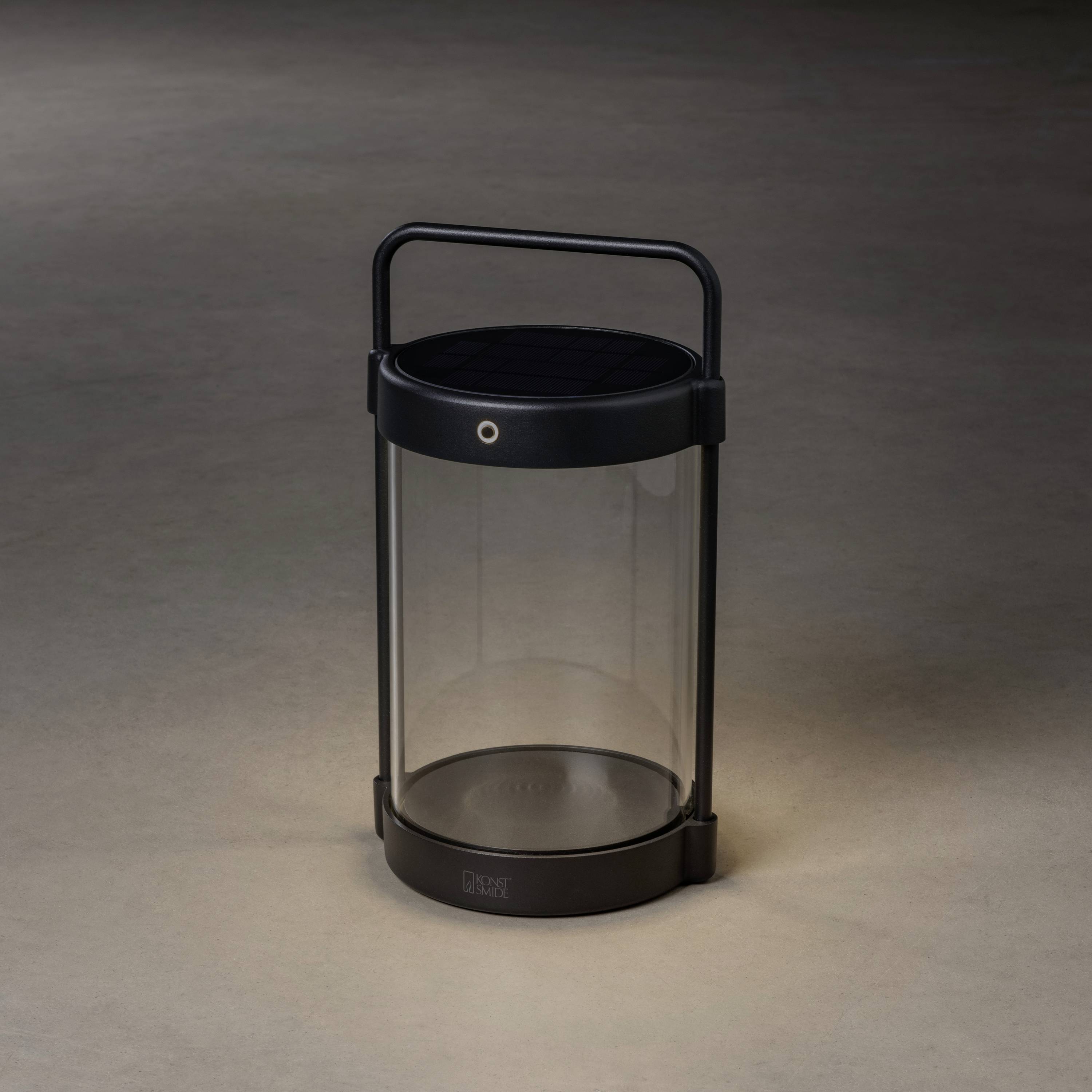 Eine tragbare, zylindrische Solar-Laterne mit dunklem Rahmen und transparentem Glas, die auf einem grauen Untergrund steht.