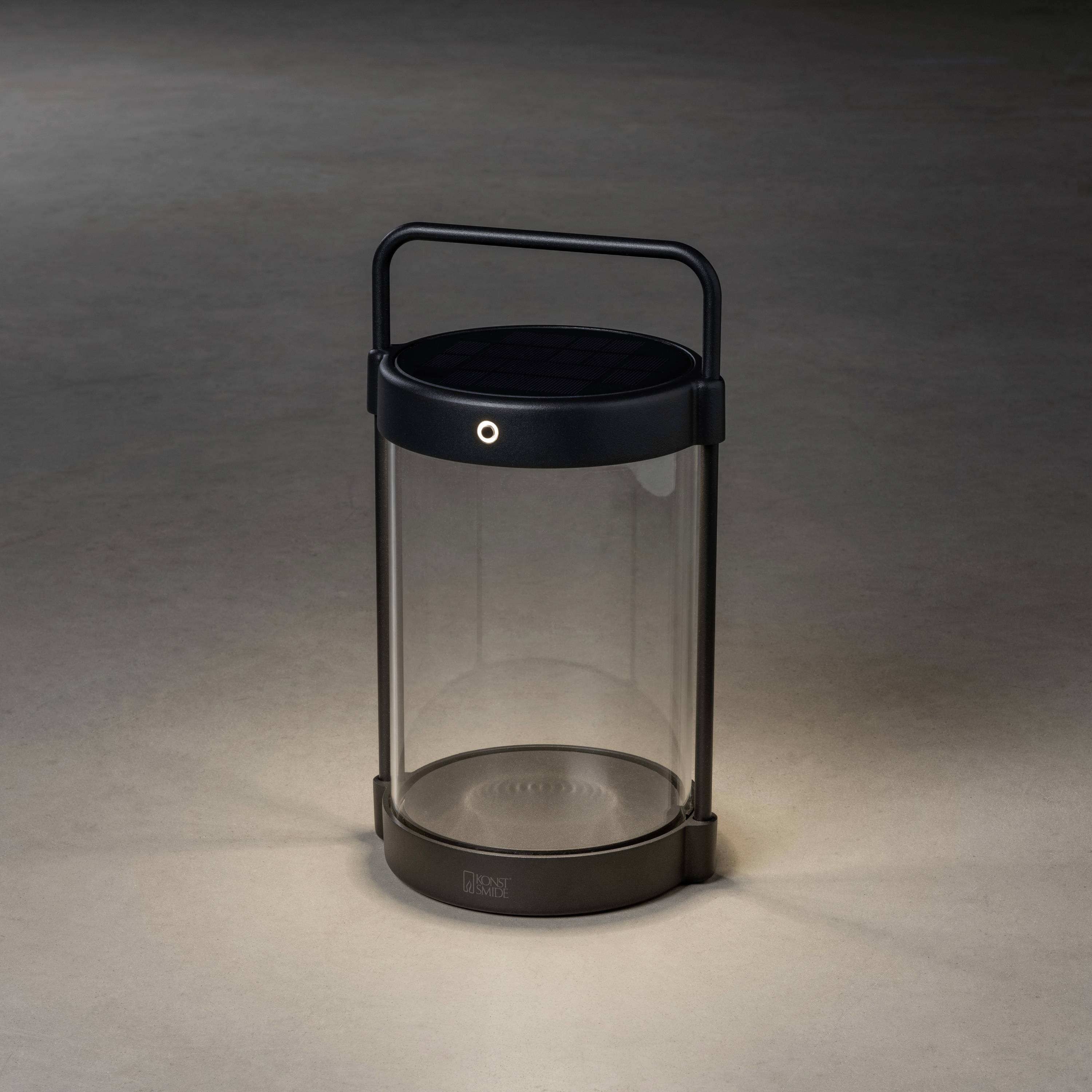 Eine tragbare Lampe mit Griff, transparentem Zylinderkörper und schwarzer Solarplatte oben, steht auf einem grauen Boden in gedämpftem Licht.