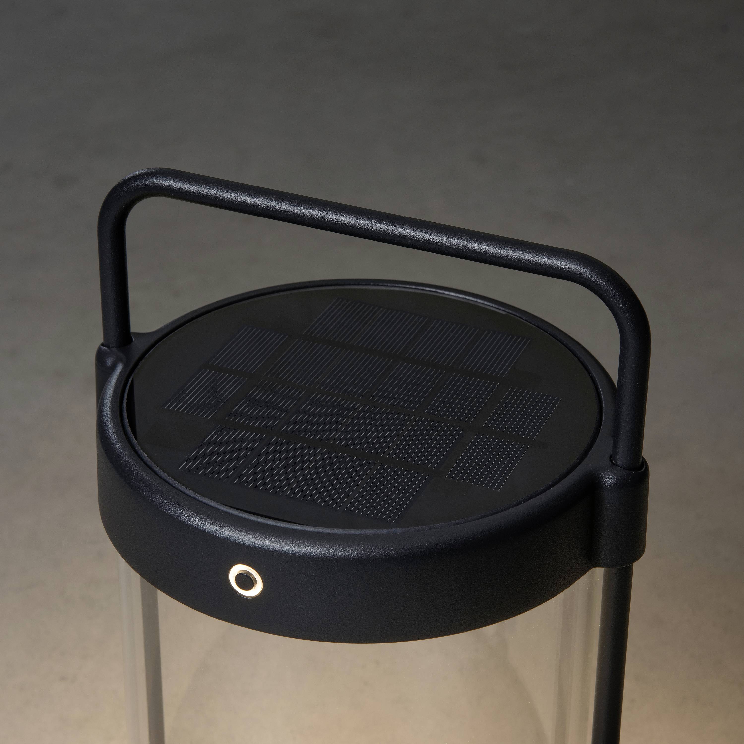 Eine tragbare Lampenlaterne mit schwarzem Griff und sichtbarem Solarpanel auf der Oberseite, steht auf einem grauen Boden.
