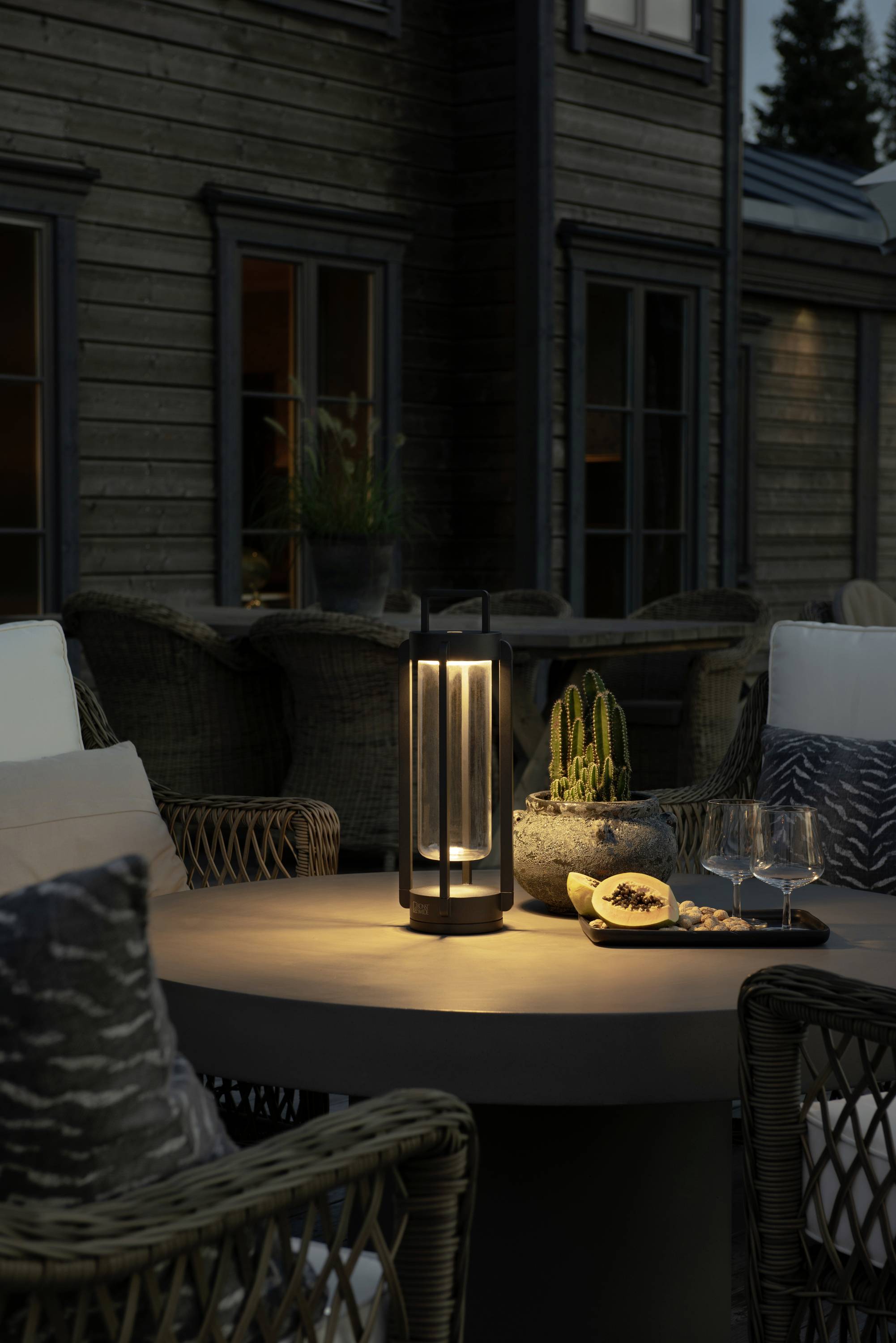 Tisch auf einer Terrasse bei Dämmerung, mit einer leuchtenden Laterne, Snacks, Weingläsern und Pflanzen darauf; Sitzgelegenheiten um den Tisch.