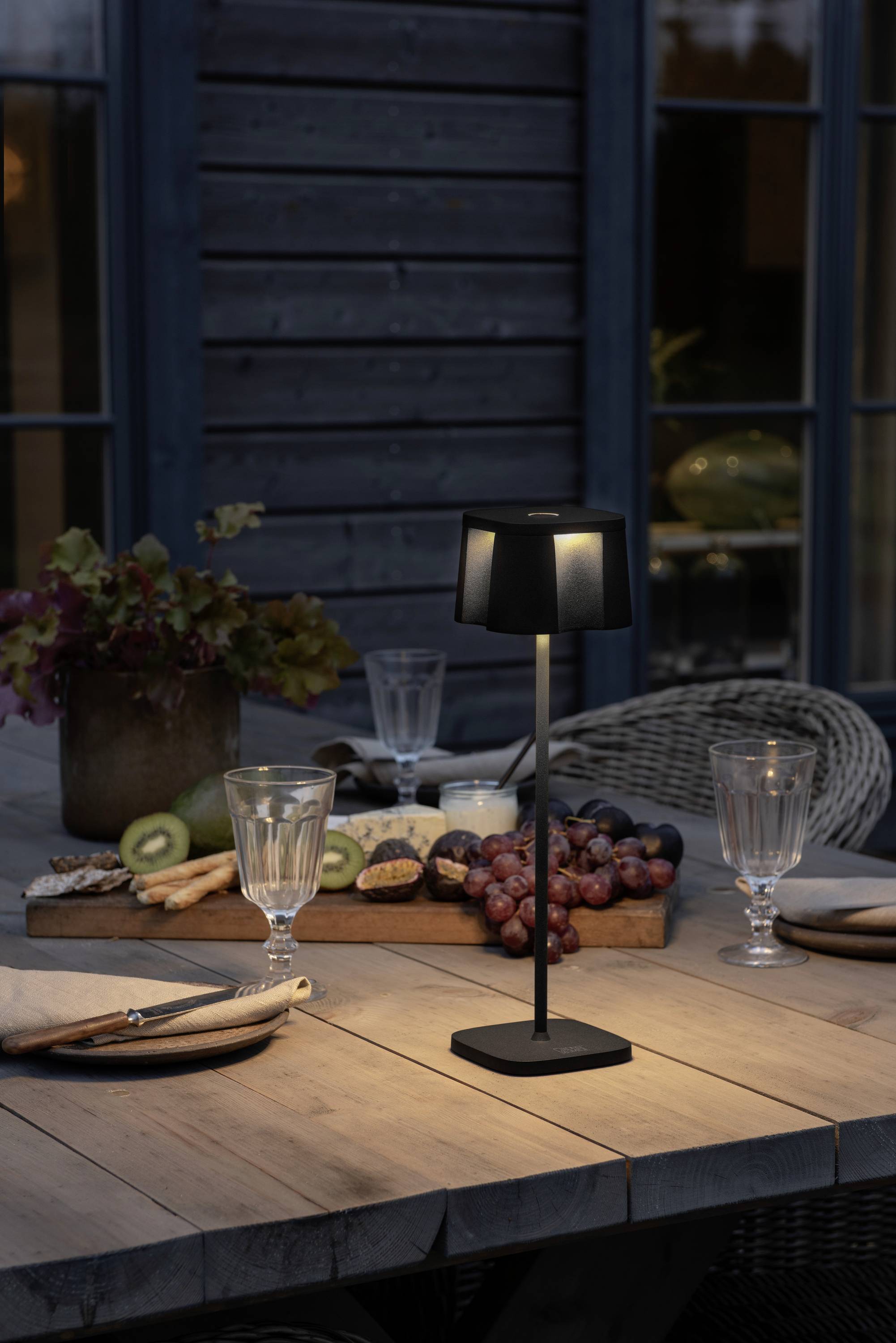 Ein gedeckter Holztisch im Freien mit einer leuchtenden Tischlampe, Gläsern, Tellern und einer Schale mit Obst und Käse.