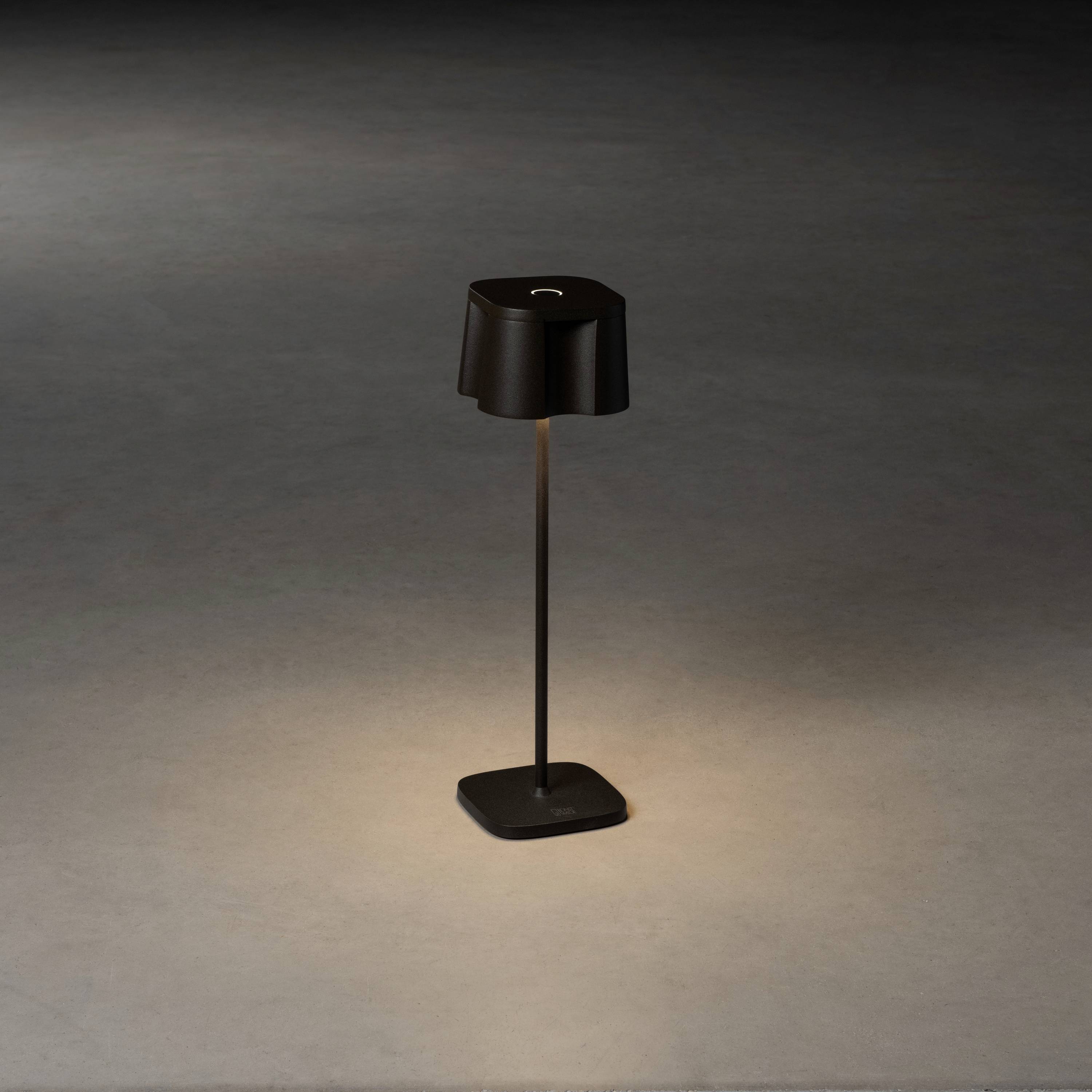 Eine schwarze Tischlampe auf einem dunklen, grauen Hintergrund. Sie ist eingeschaltet und wirft einen sanften Lichtkegel nach unten.