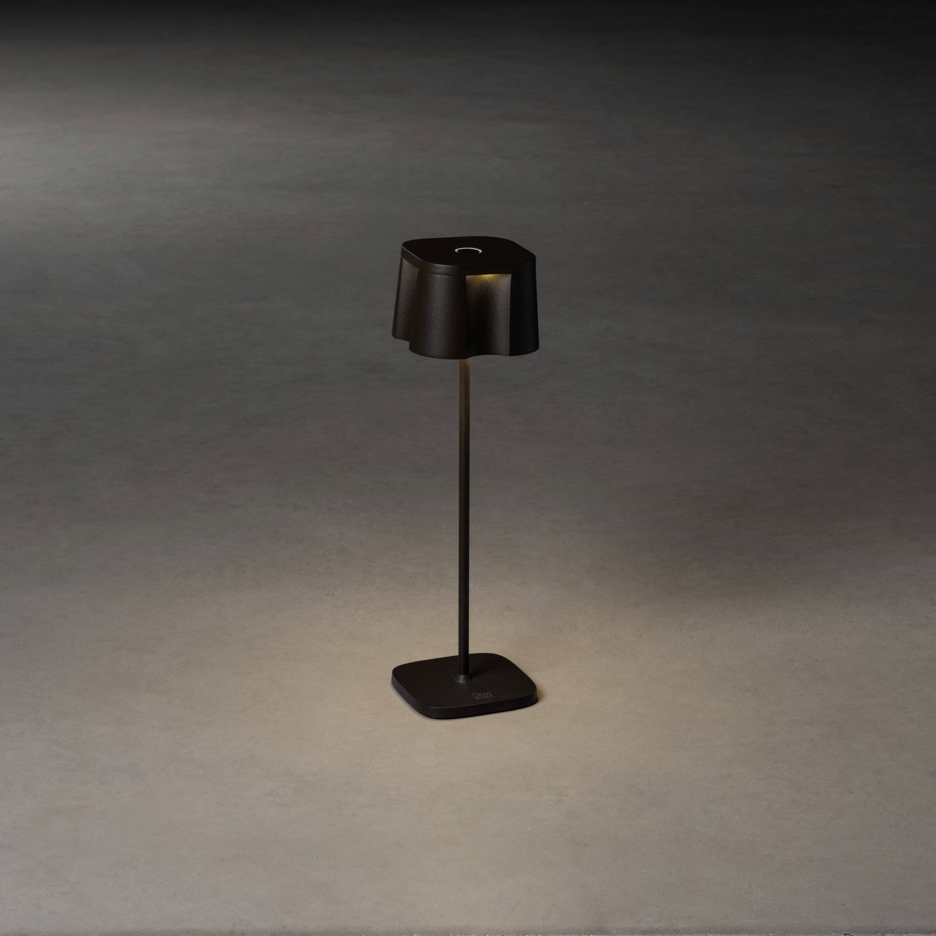 Eine schwarze Stehlampe mit schlichtem Design steht auf einem grauen Boden. Sie strahlt Licht nach unten und erzeugt einen kleinen Lichtkegel.