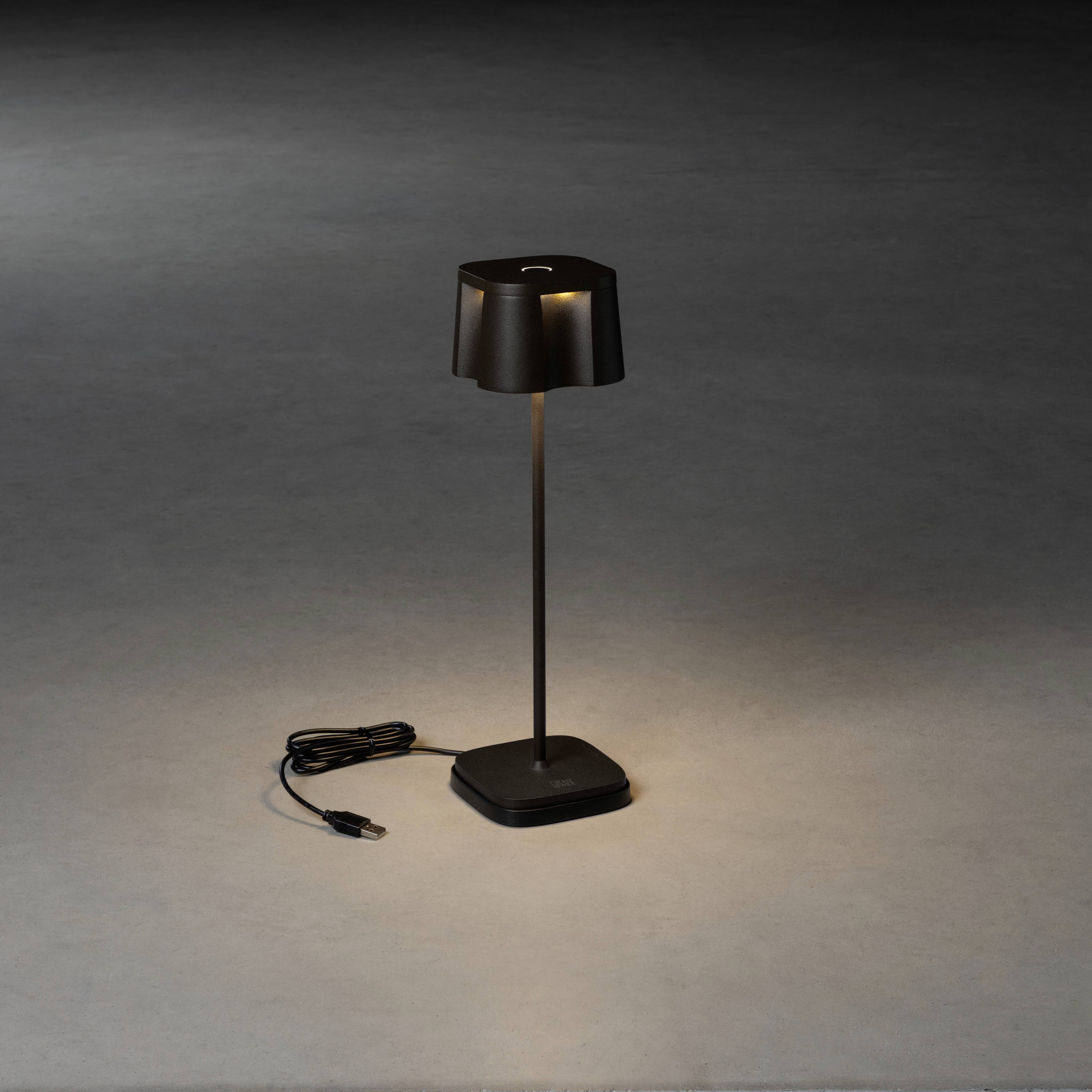 Schwarze Tischlampe mit schmalem Ständer und quadratischem Lampenschirm, eingeschaltet, wirft Licht auf dunkler Oberfläche. Kabel sichtbar.