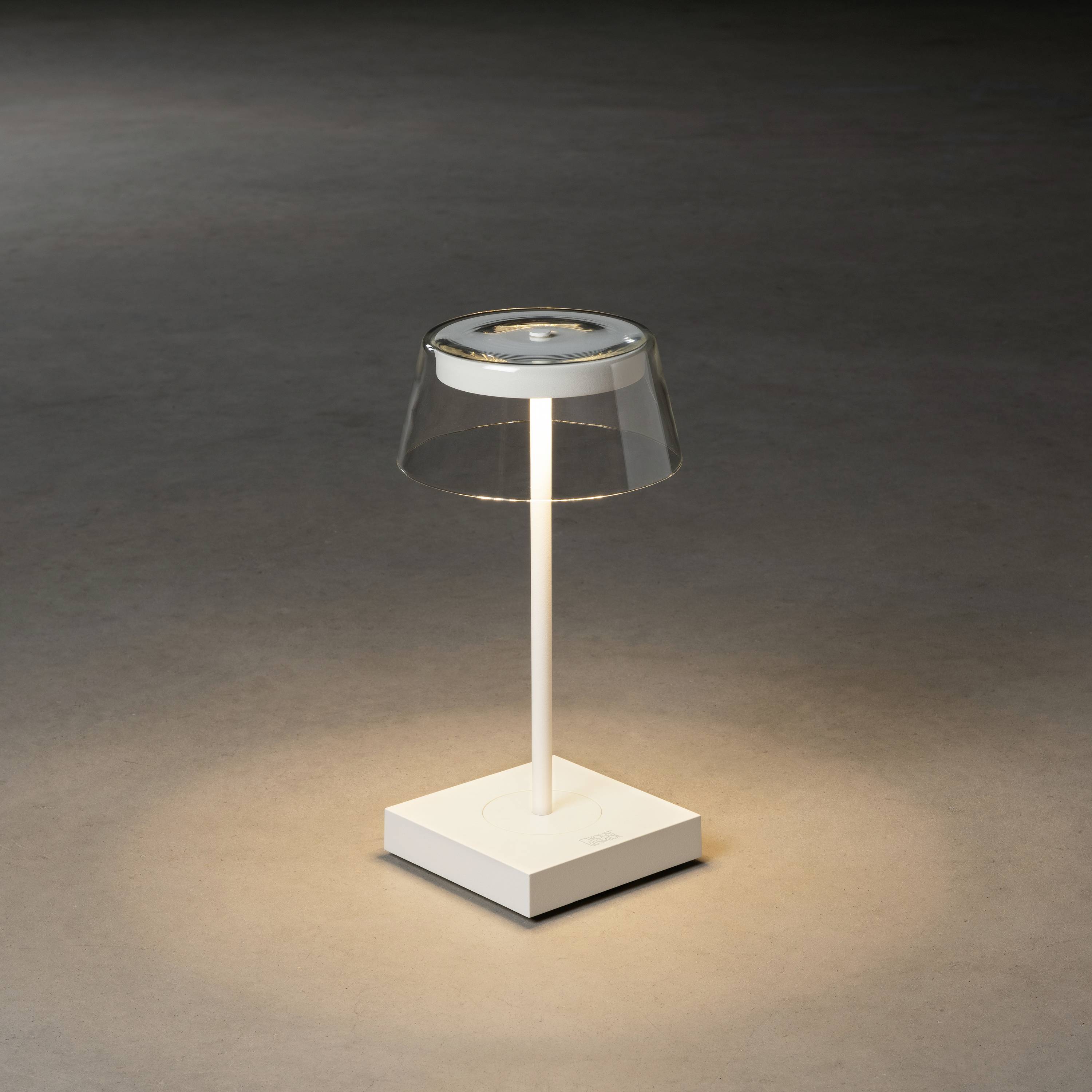 Eine moderne Tischlampe mit transparentem Schirm wirft weiches Licht auf den Boden. Sie steht auf einem weißen rechteckigen Sockel.
