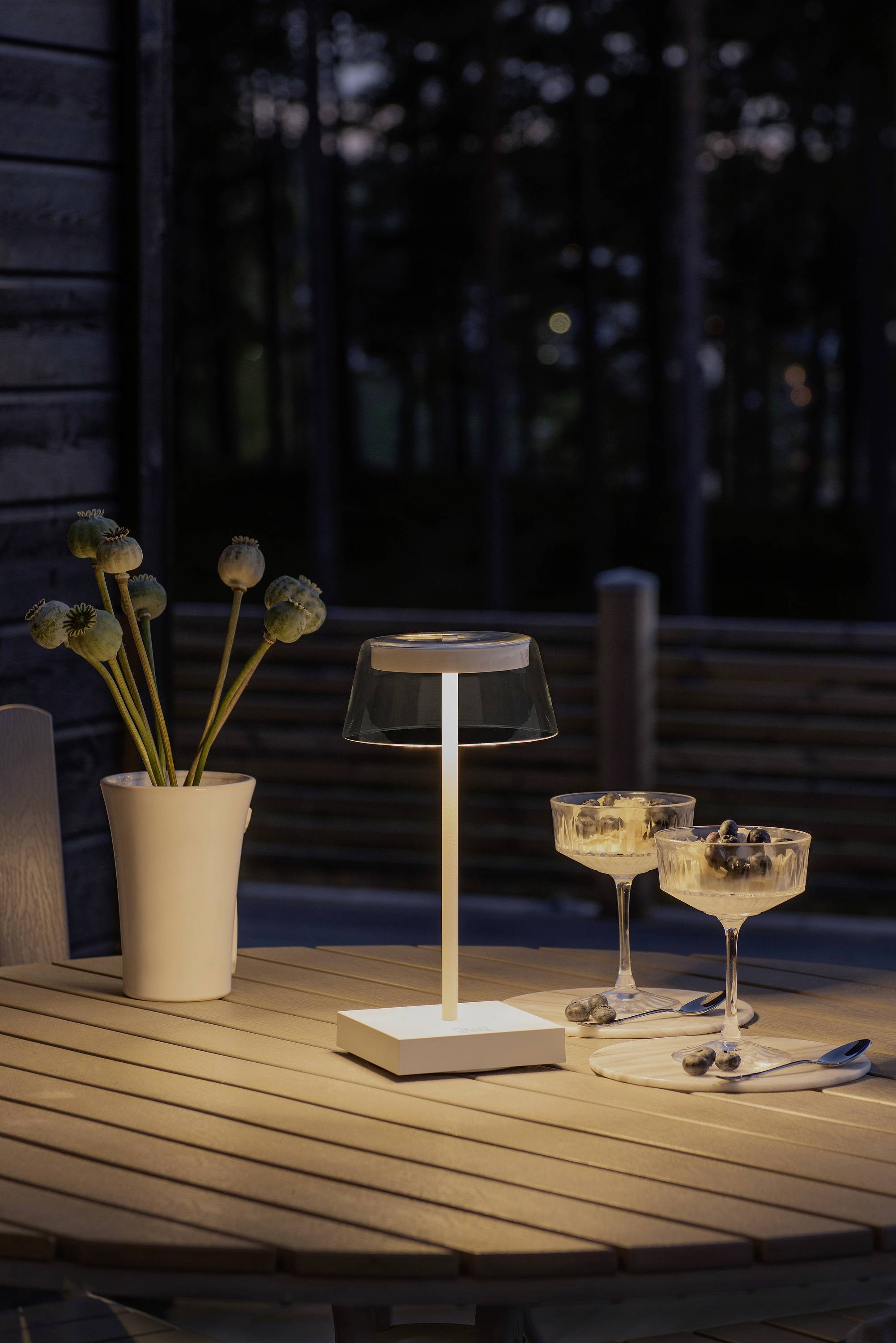 Eine Tischlampe leuchtet auf einem Holztisch im Freien, neben zwei Gläsern mit Dessert und einer Vase mit Blumen; es ist abends.