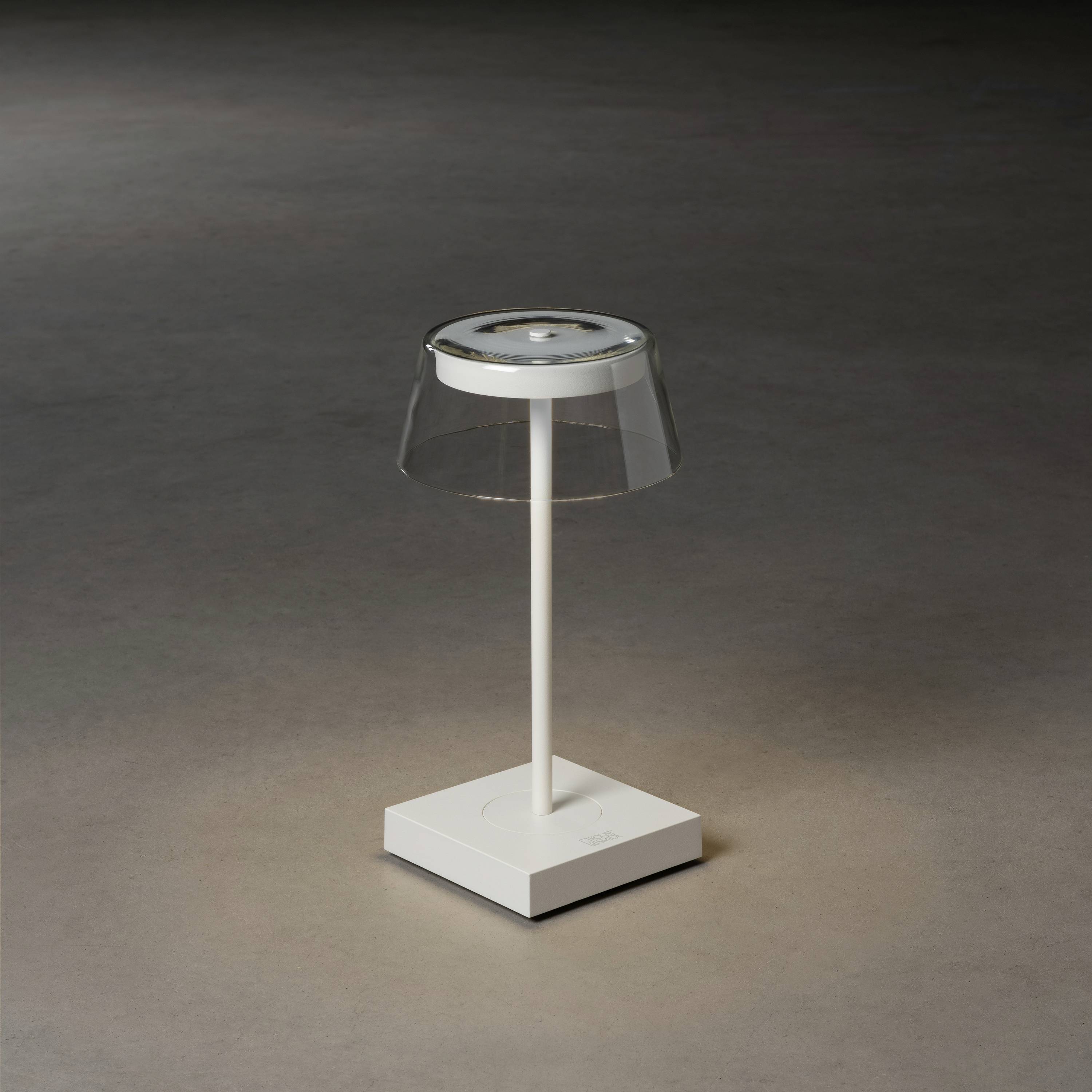 Eine moderne Tischlampe mit transparentem Schirm auf quadratischem Sockel in minimalistischem Design, leuchtet sanft auf dunklem Hintergrund.