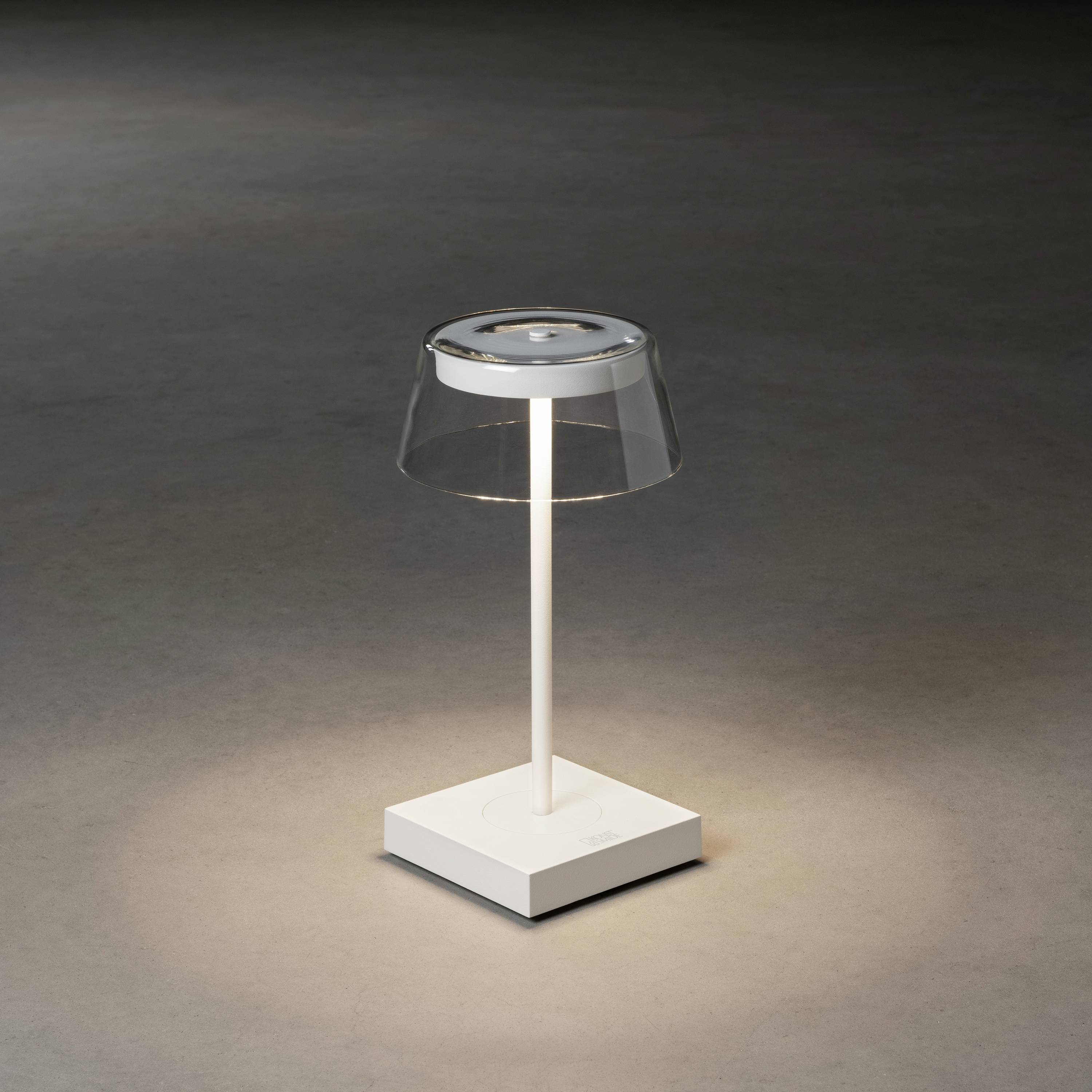 Eine moderne, minimalistische Tischlampe mit transparentem Schirm, stehend auf einem quadratischen Sockel in einem dunklen Raum.