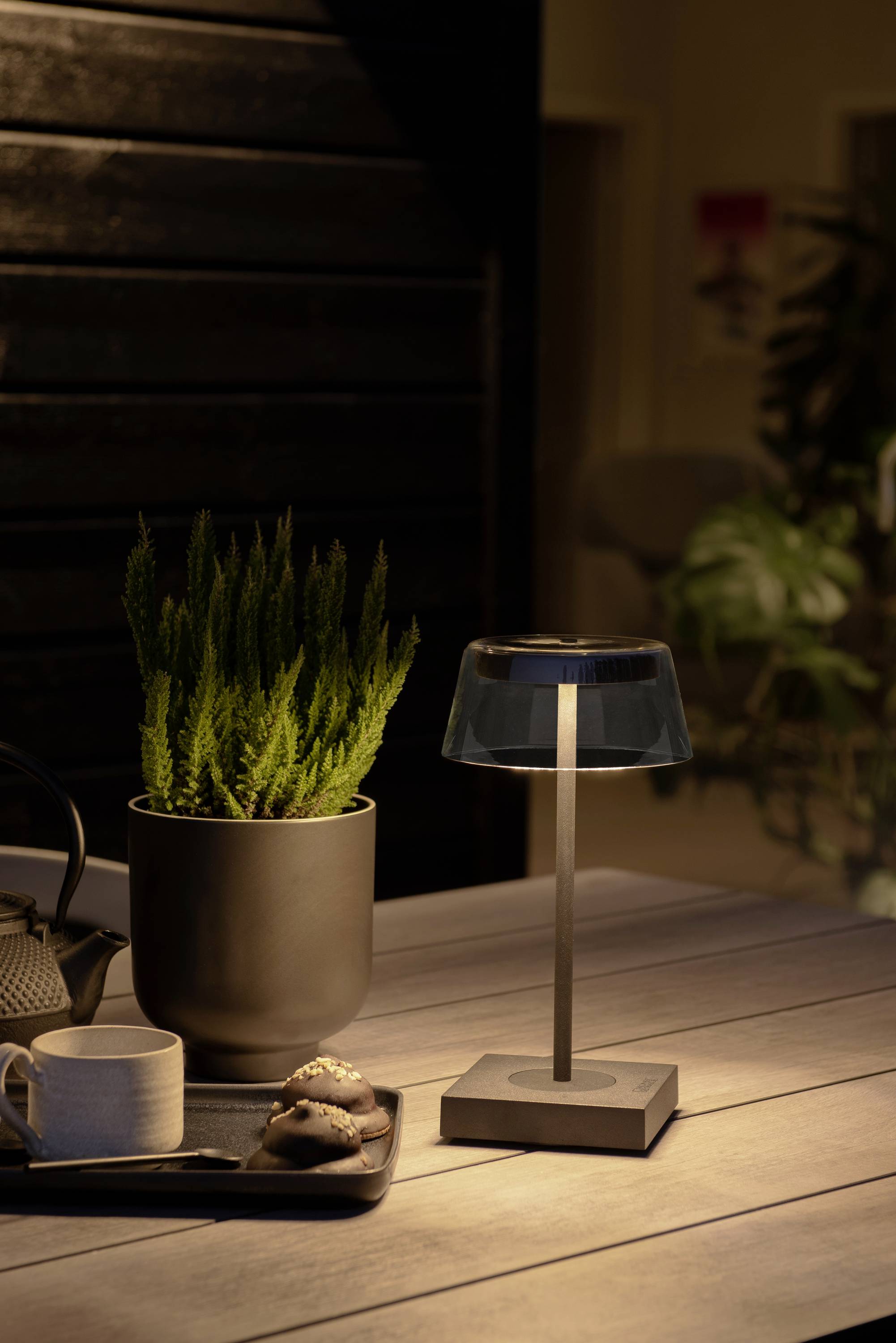 Ein beleuchteter Tisch mit einer kleinen Lampe, einem Blumentopf mit grünem Kraut, einer Teekanne, einer Tasse und Keksen auf einem Tablett.