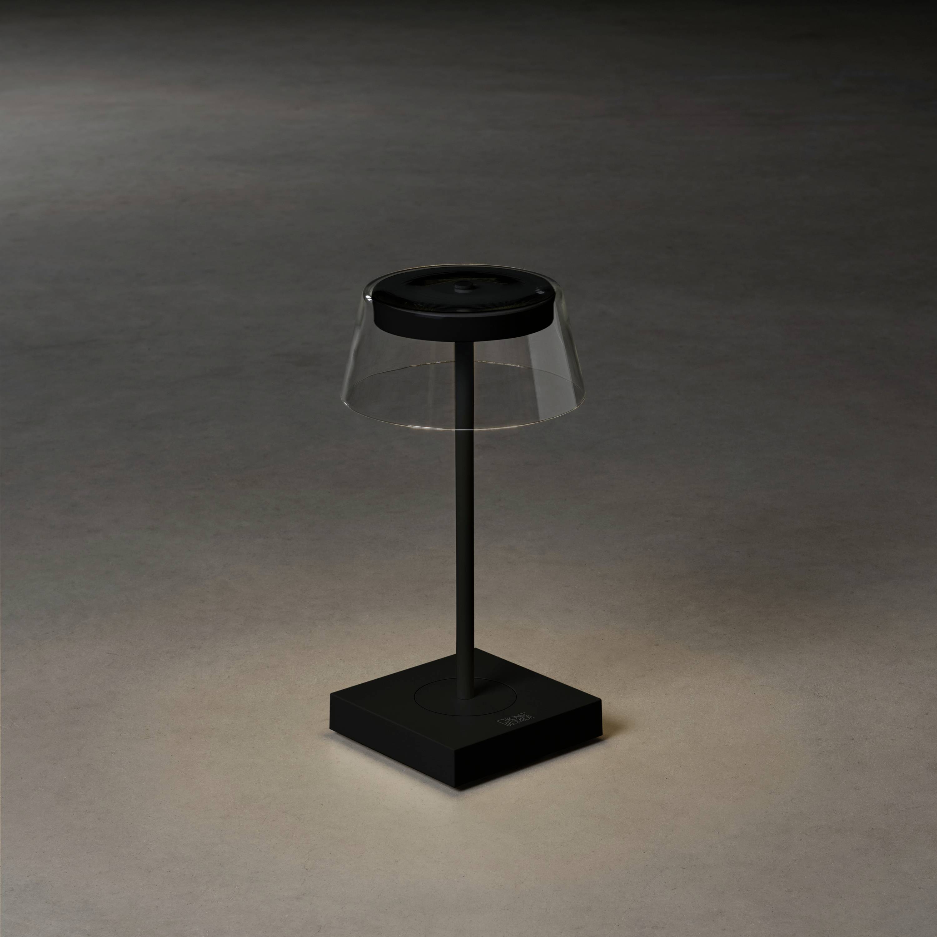 Eine moderne Tischlampe mit transparentem Schirm und schwarzem Sockel steht in einem dunklen Raum und beleuchtet leicht den Boden.