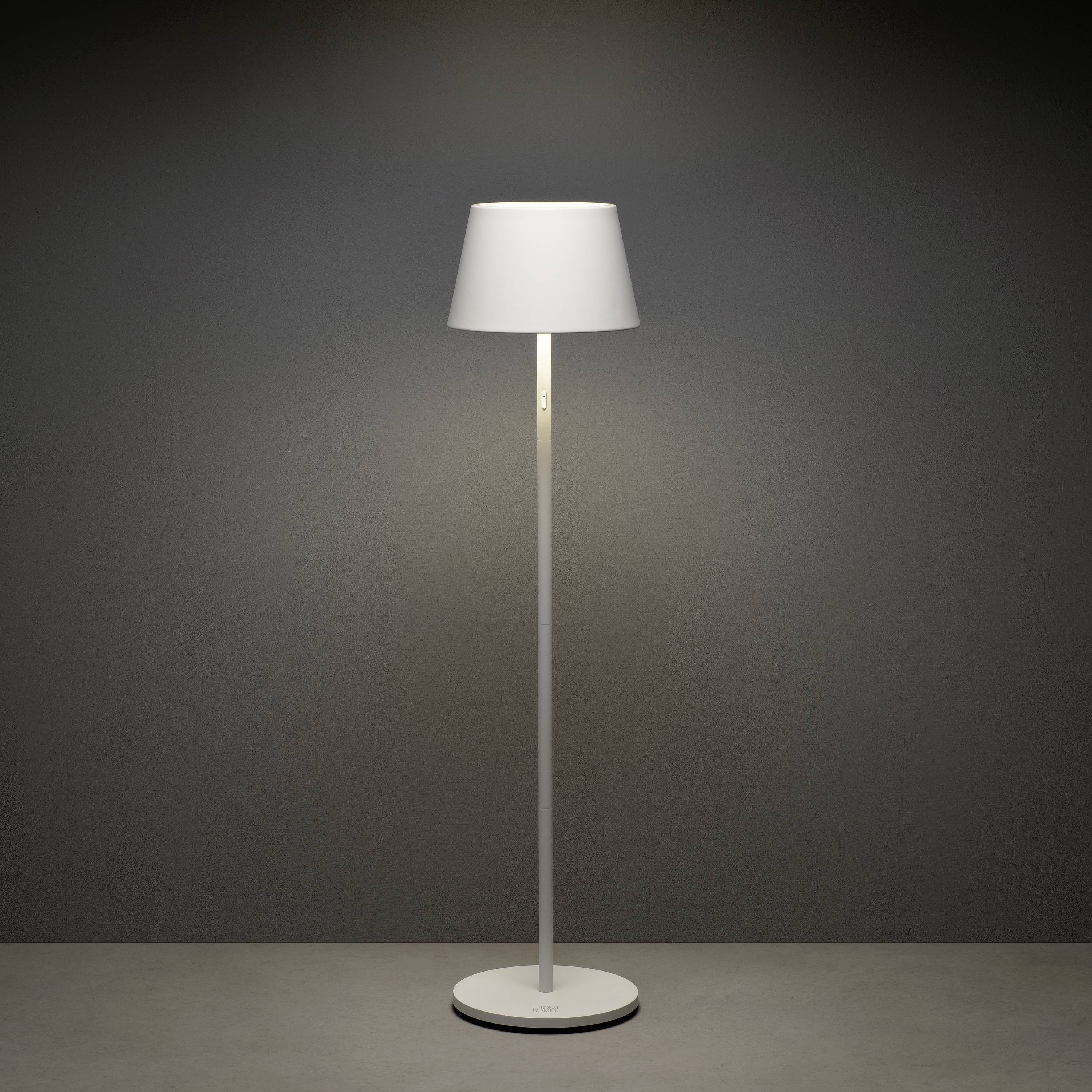 Eine schlichte, moderne Stehlampe mit weißem Schirm steht in einem dunkelgrauen Raum. Die Lampe ist eingeschaltet und strahlt Licht aus.