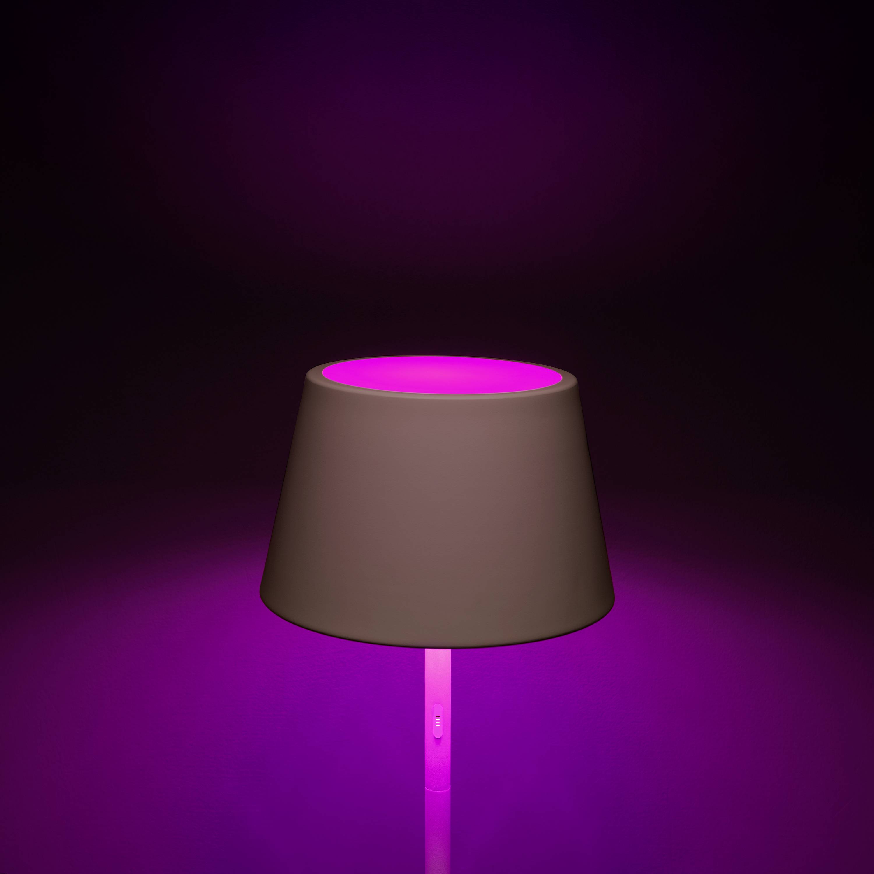 Eine Tischlampe mit einem braunen Schirm leuchtet in kräftigem Pink vor einem dunklen Hintergrund.