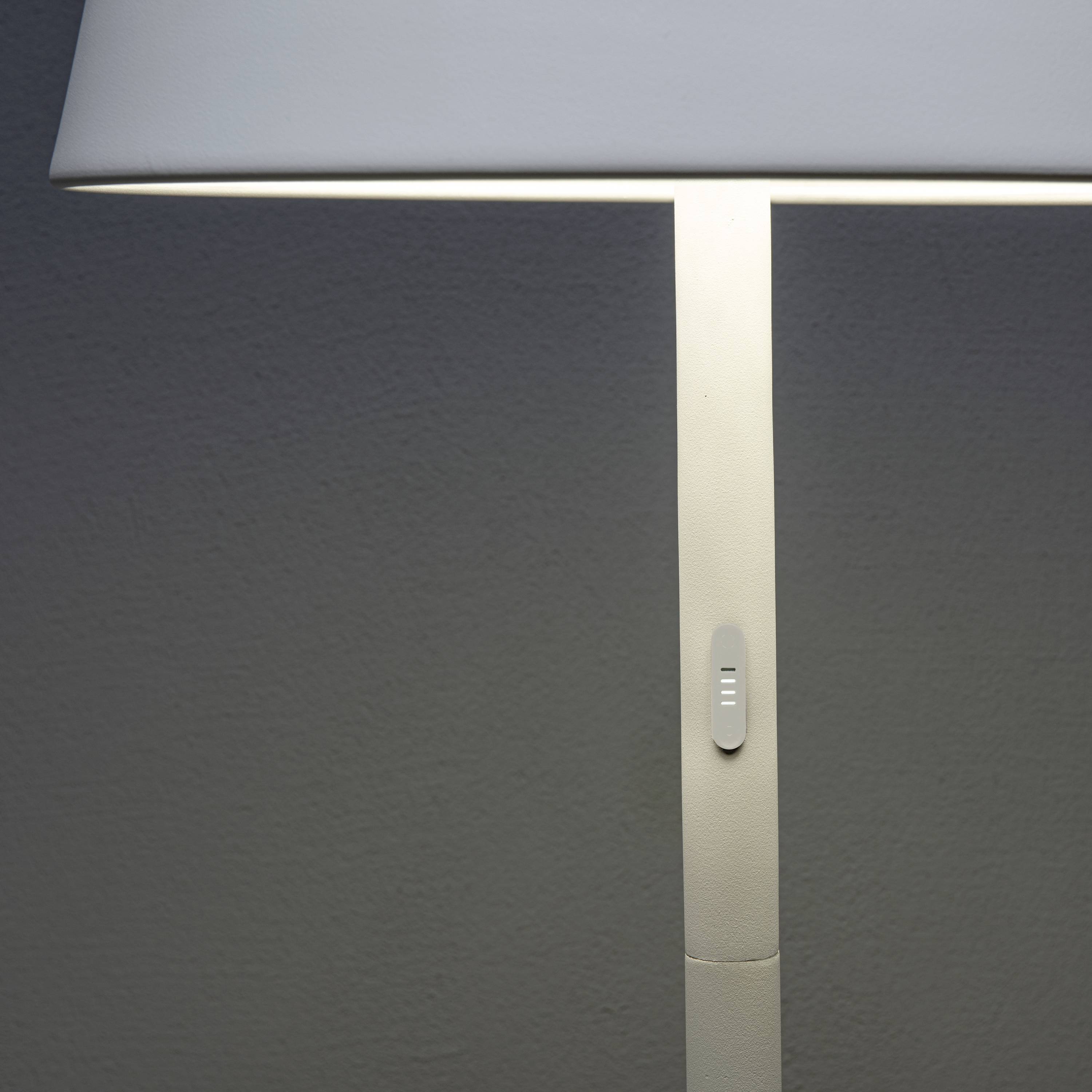 Eine schlichte, moderne Stehlampe mit weißem Lampenschirm leuchtet in einem Raum. Der Schalter befindet sich am Ständer der Lampe.