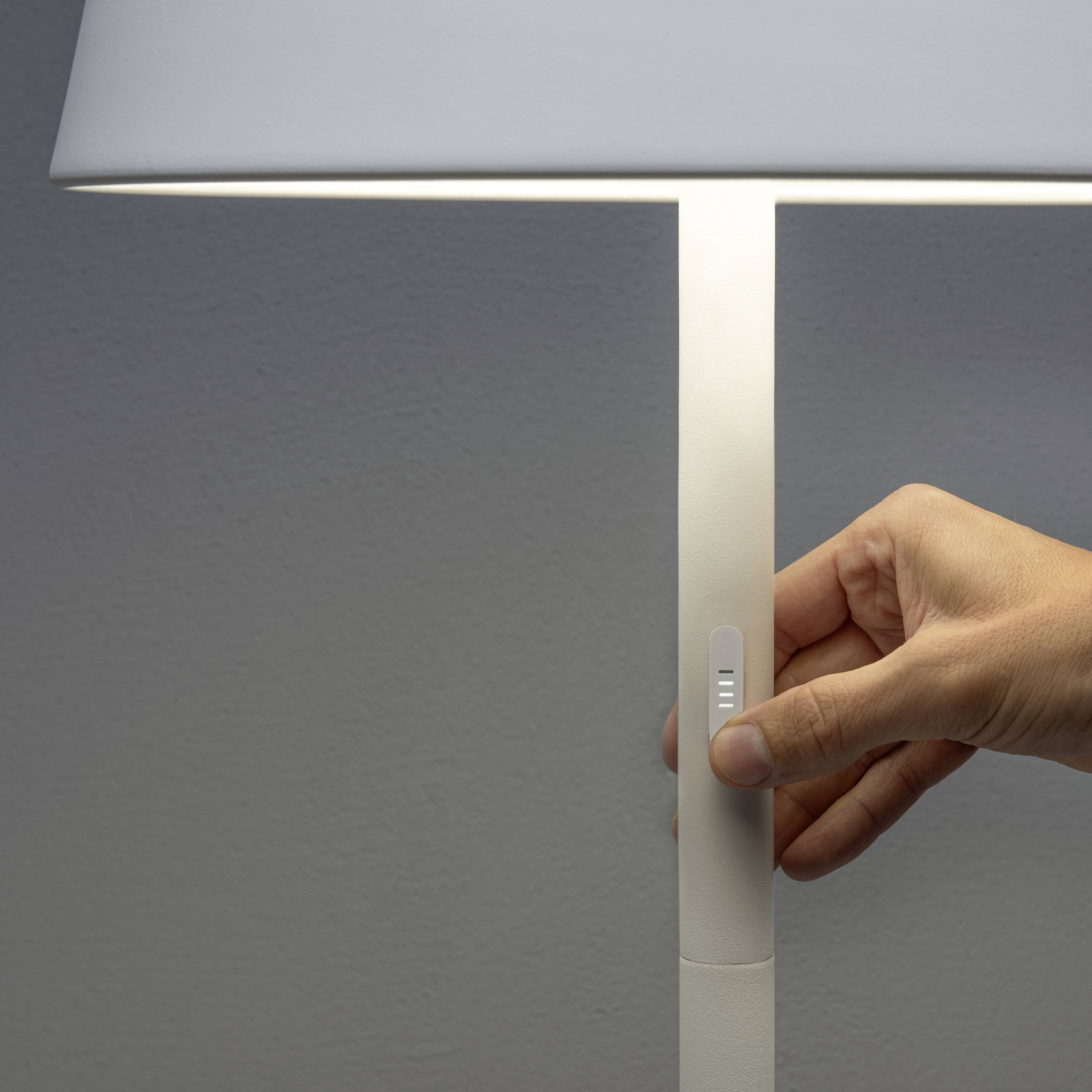 Eine Hand berührt den Schalter einer modernen, minimalistisch gestalteten Tischlampe mit einem weißen Lampenschirm.