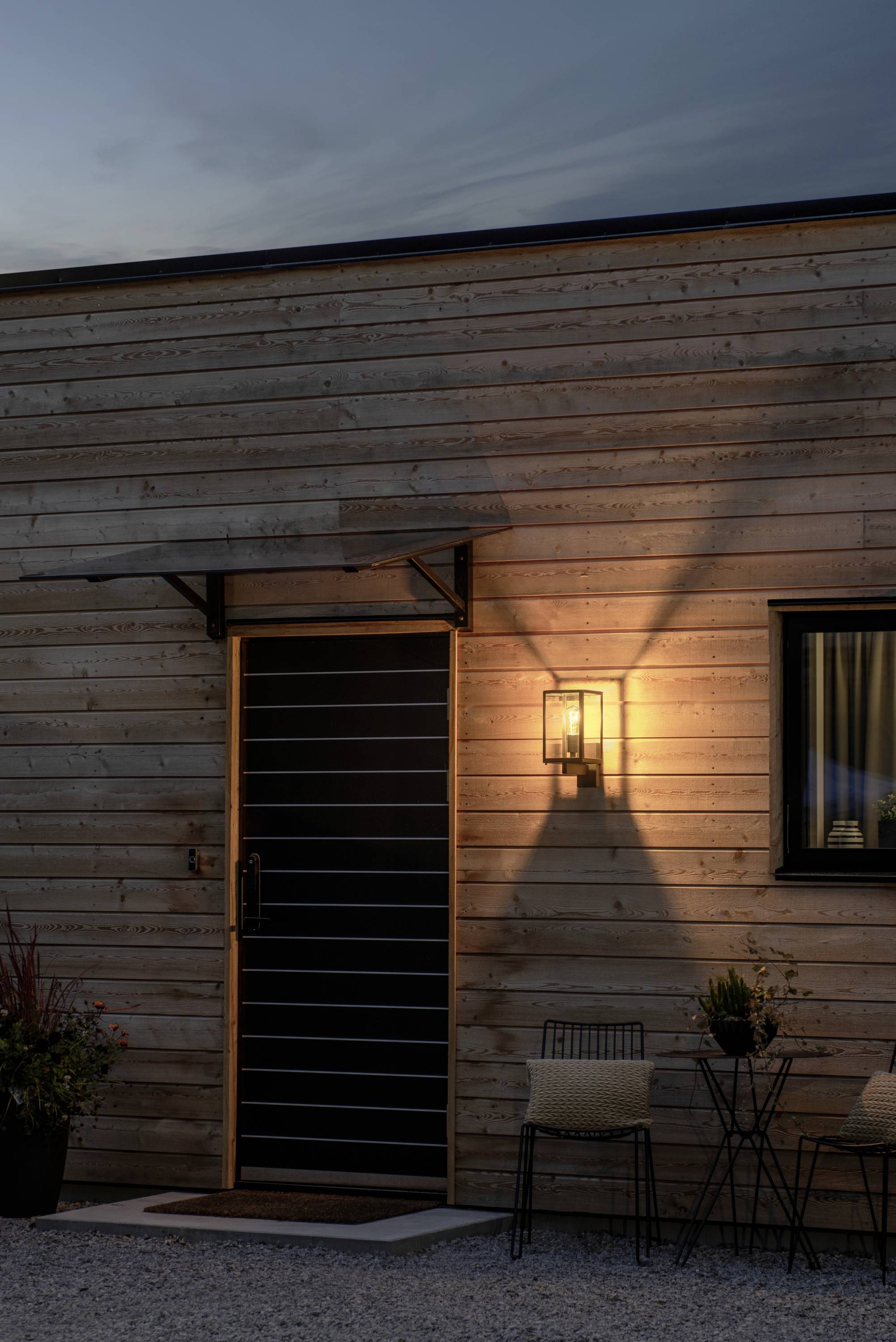 Ein modernes Holzhaus mit einer Lampe, die warmes Licht auf die Fassade und den Boden wirft. Neben der Tür steht ein kleiner Tisch mit zwei Stühlen.