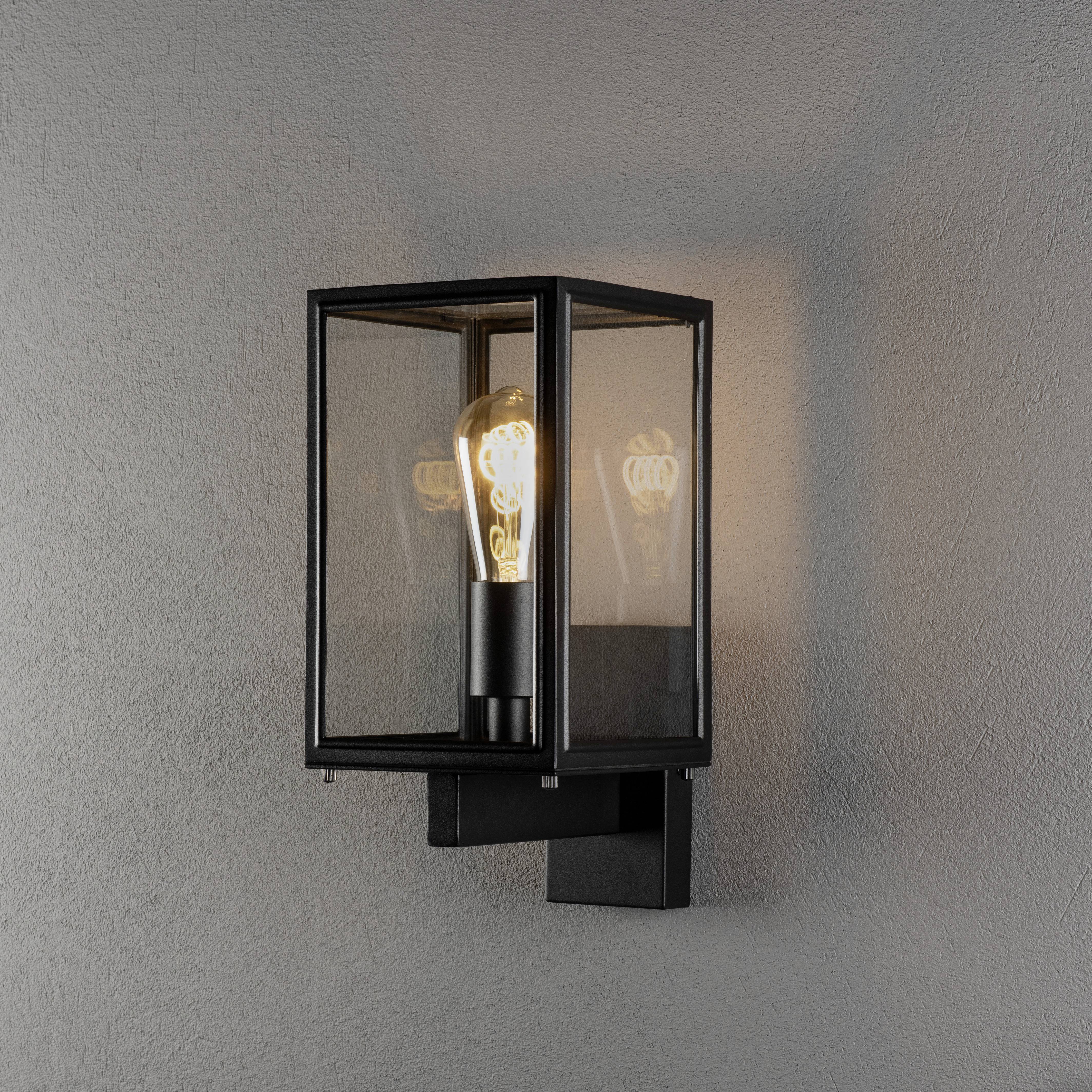 Wandleuchte mit schwarzem Rahmen und Glaseinsatz, beleuchtet von einer sichtbaren Glühbirne. Sie wirft weiches Licht auf eine graue Wand.