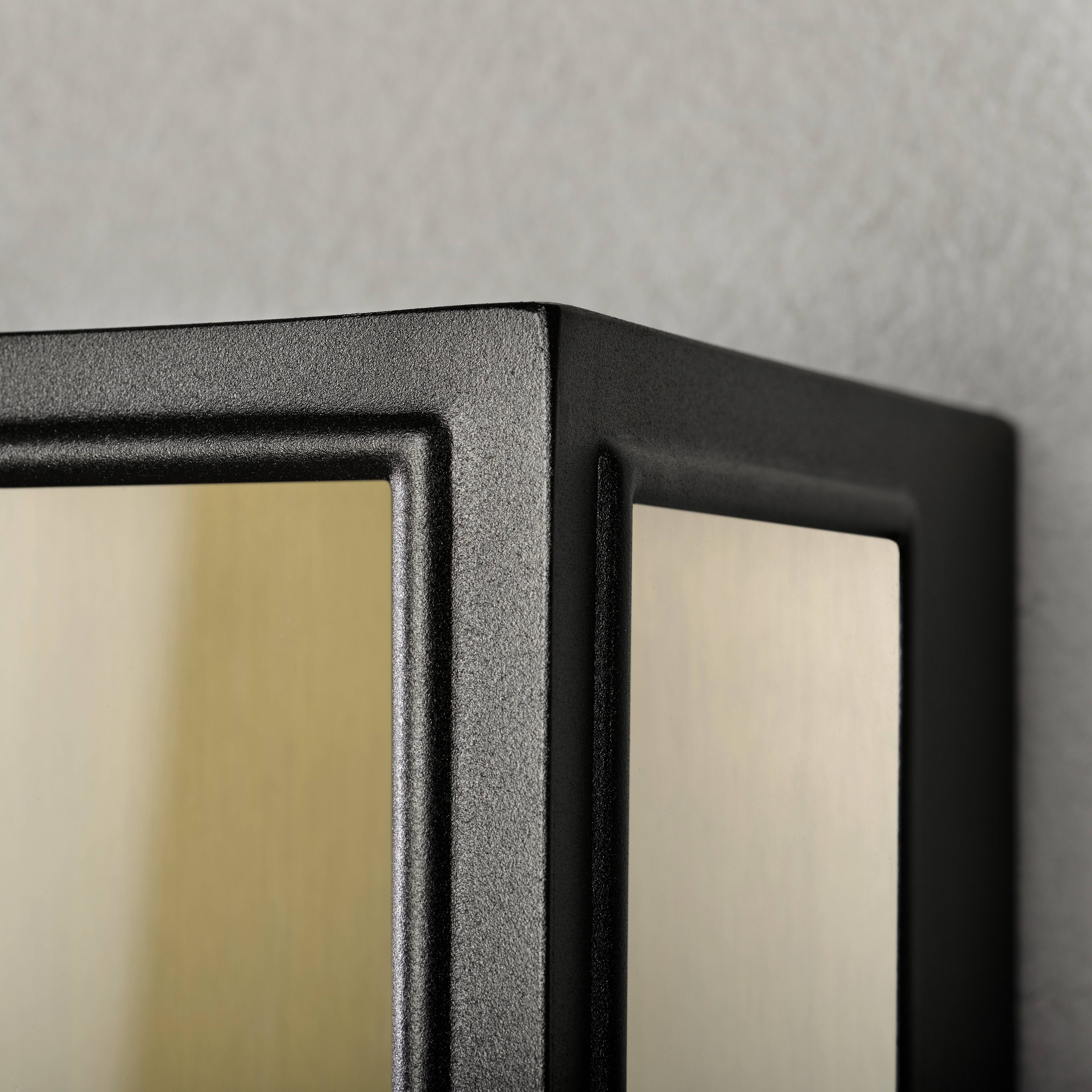 Eine moderne, schwarze Wandleuchte mit rechteckigem Rahmen ist an einer grauen Wand montiert. Sie strahlt warmes, harmonisches Licht aus.