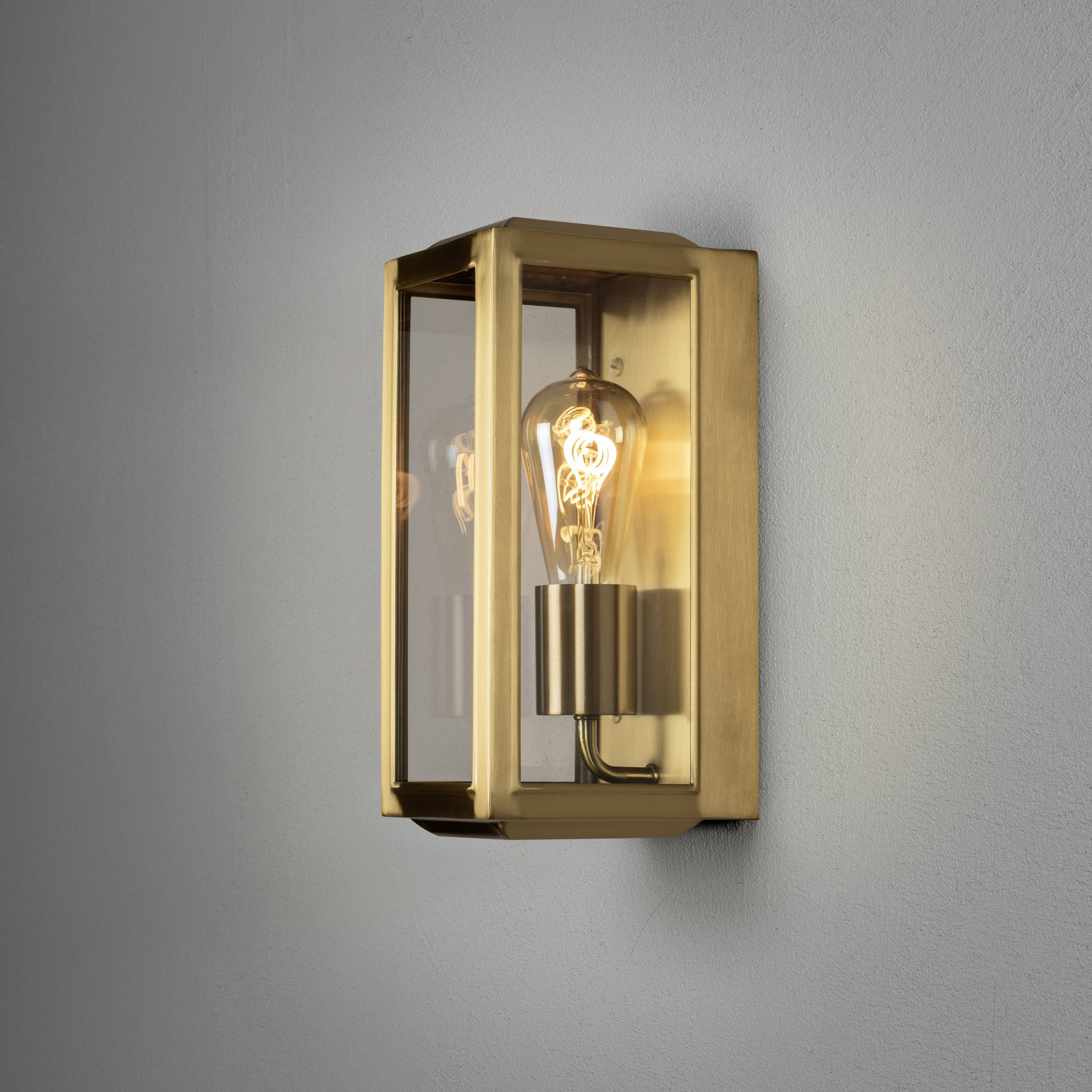 An der Wand montierte goldene Wandleuchte mit sichtbarer Glühbirne in einem rechteckigen, transparenten Gehäuse.