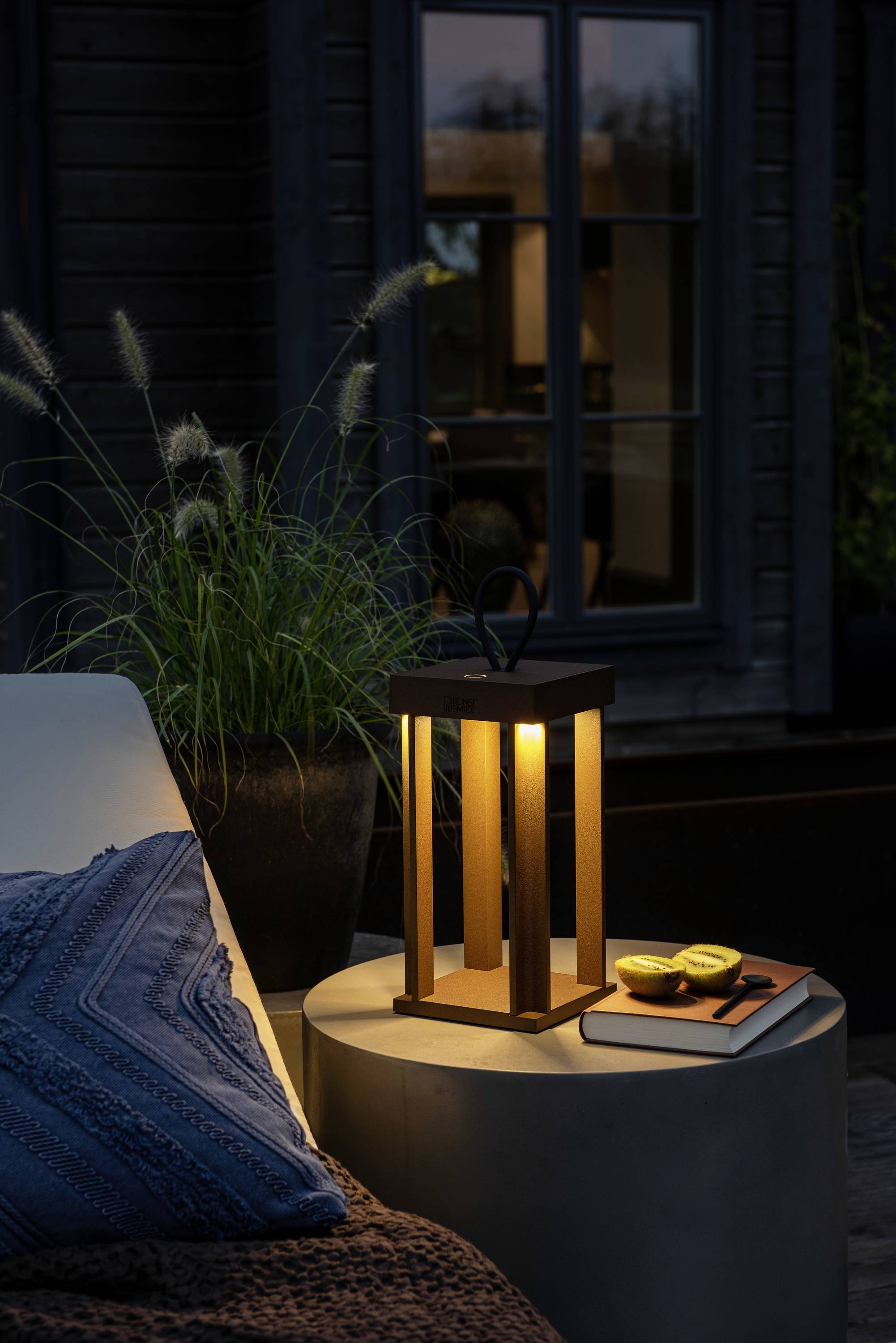 Eine Laterne auf einem Tisch beleuchtet eine Terrasse bei Dämmerung. Daneben liegen zwei Zitronen und ein Buch. Ein gemütliches Ambiente.