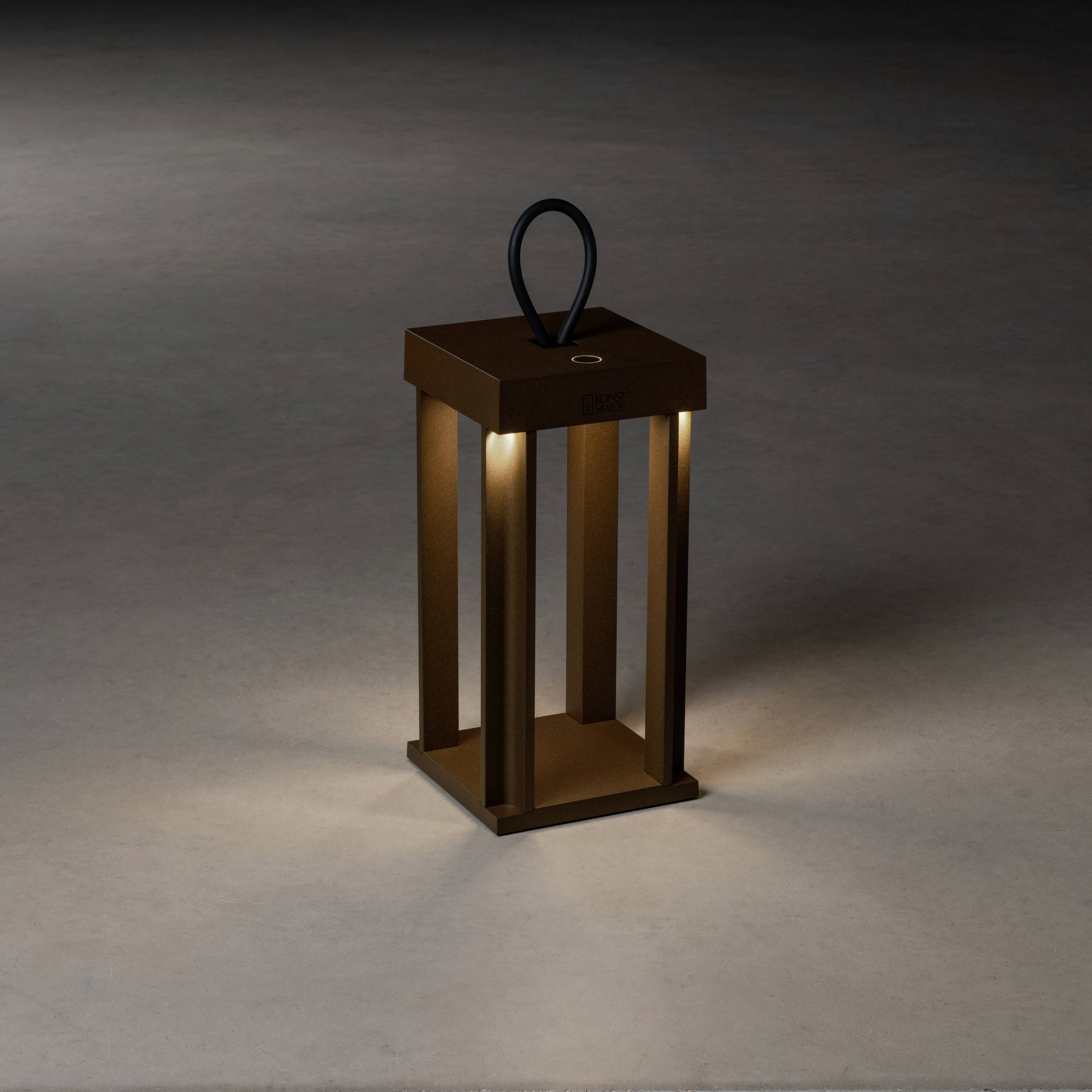 Eine moderne, quadratische, bronzefarbene Tischleuchte mit Griff und vier offenen Seiten, die weiches Licht auf den Boden wirft.