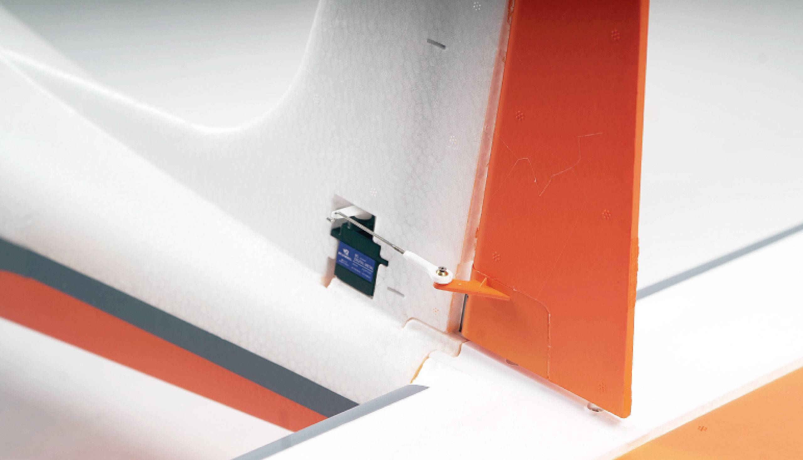 Nahaufnahme eines Modellflugzeug-Heckflügels mit orange-weißer Farbgebung und einer Steuerkomponente am Ruder befestigt.