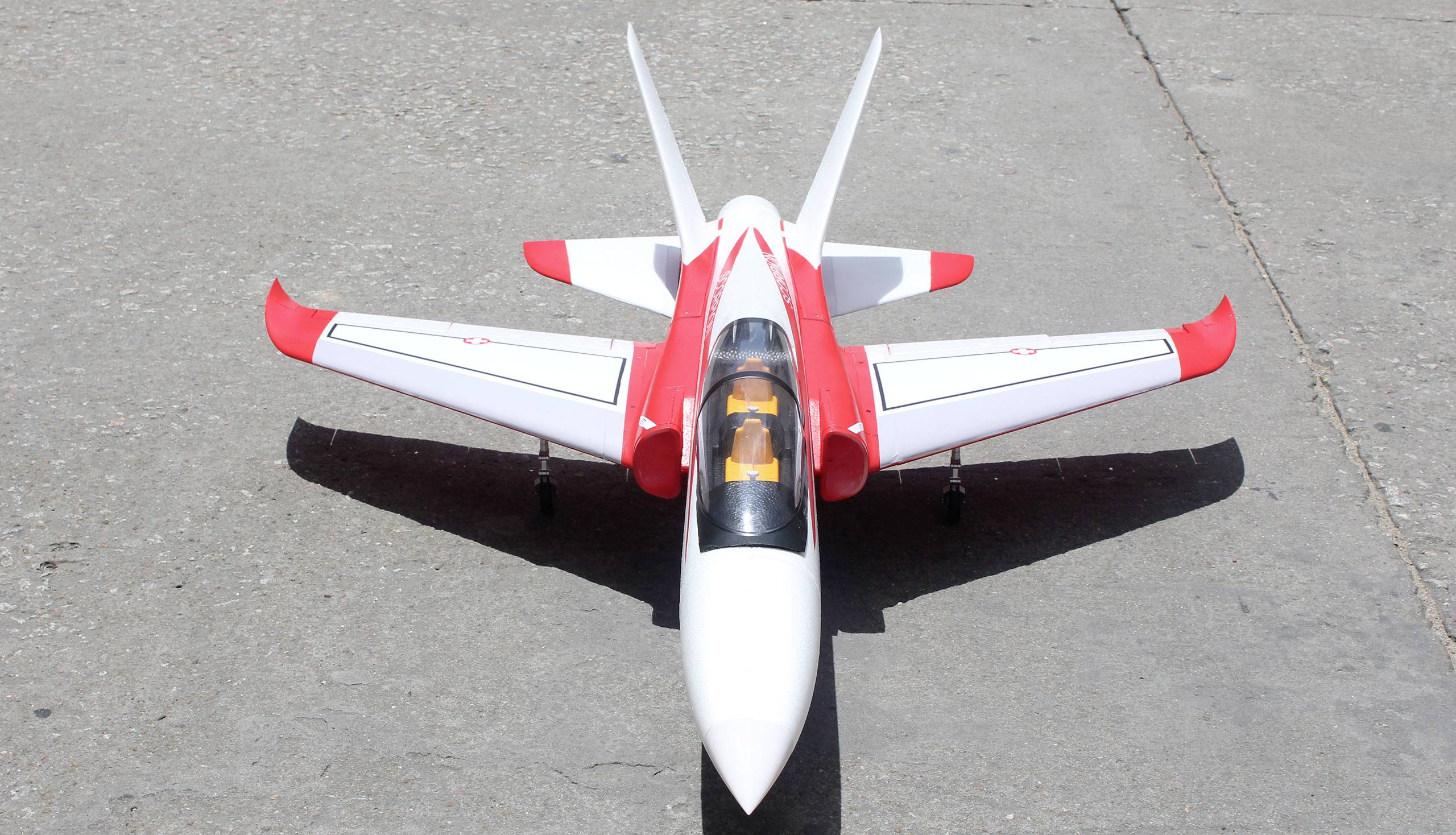 Ein weiß-rotes Modellflugzeug auf einer Betonoberfläche, von vorne fotografiert. Zwei Pilotenfiguren sind im Cockpit zu sehen.