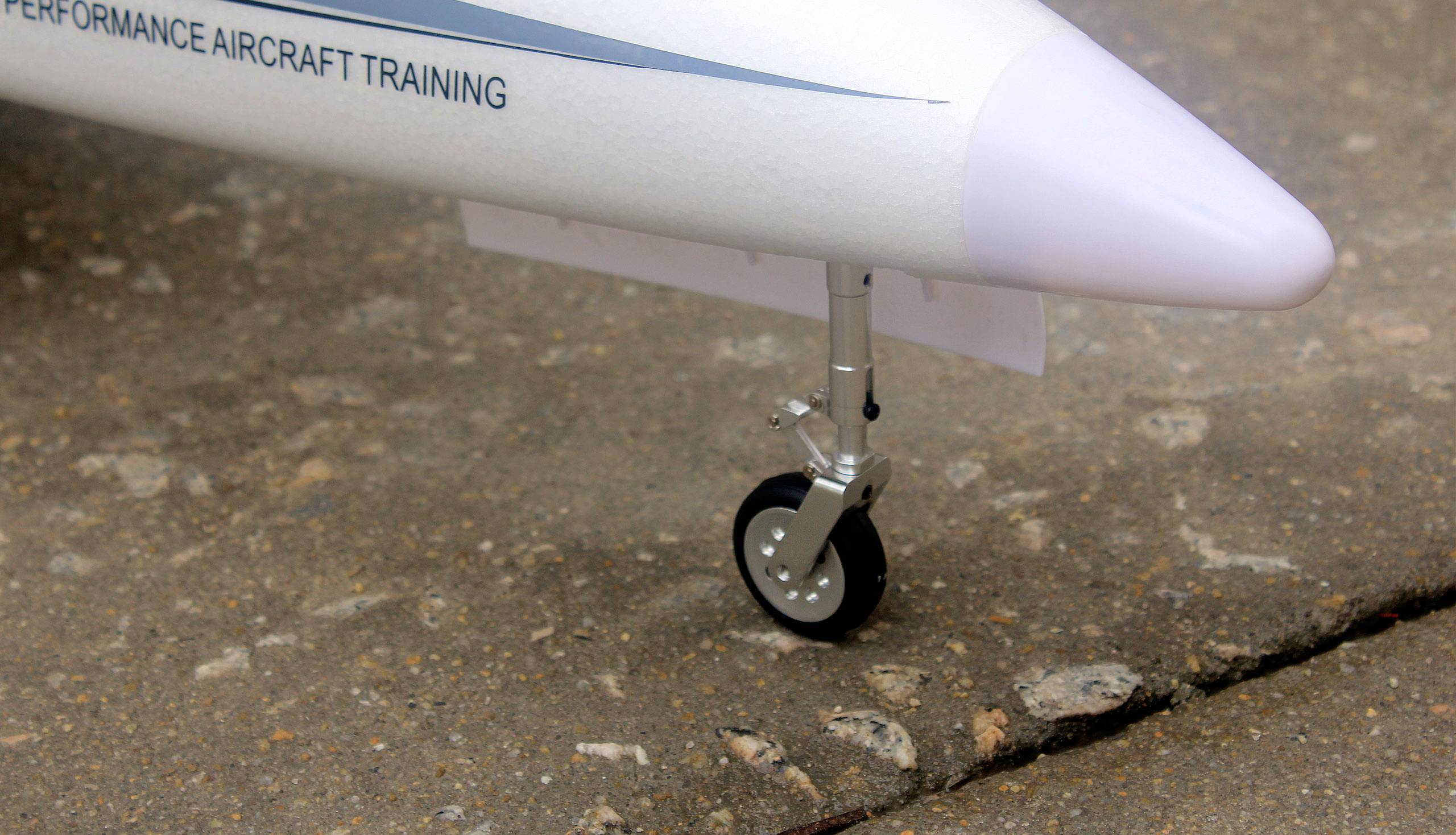 Ein kleineres Vorderfahrwerk eines Flugzeugmodells auf einer Betonfläche, mit Aufschrift 'Performance Aircraft Training'.