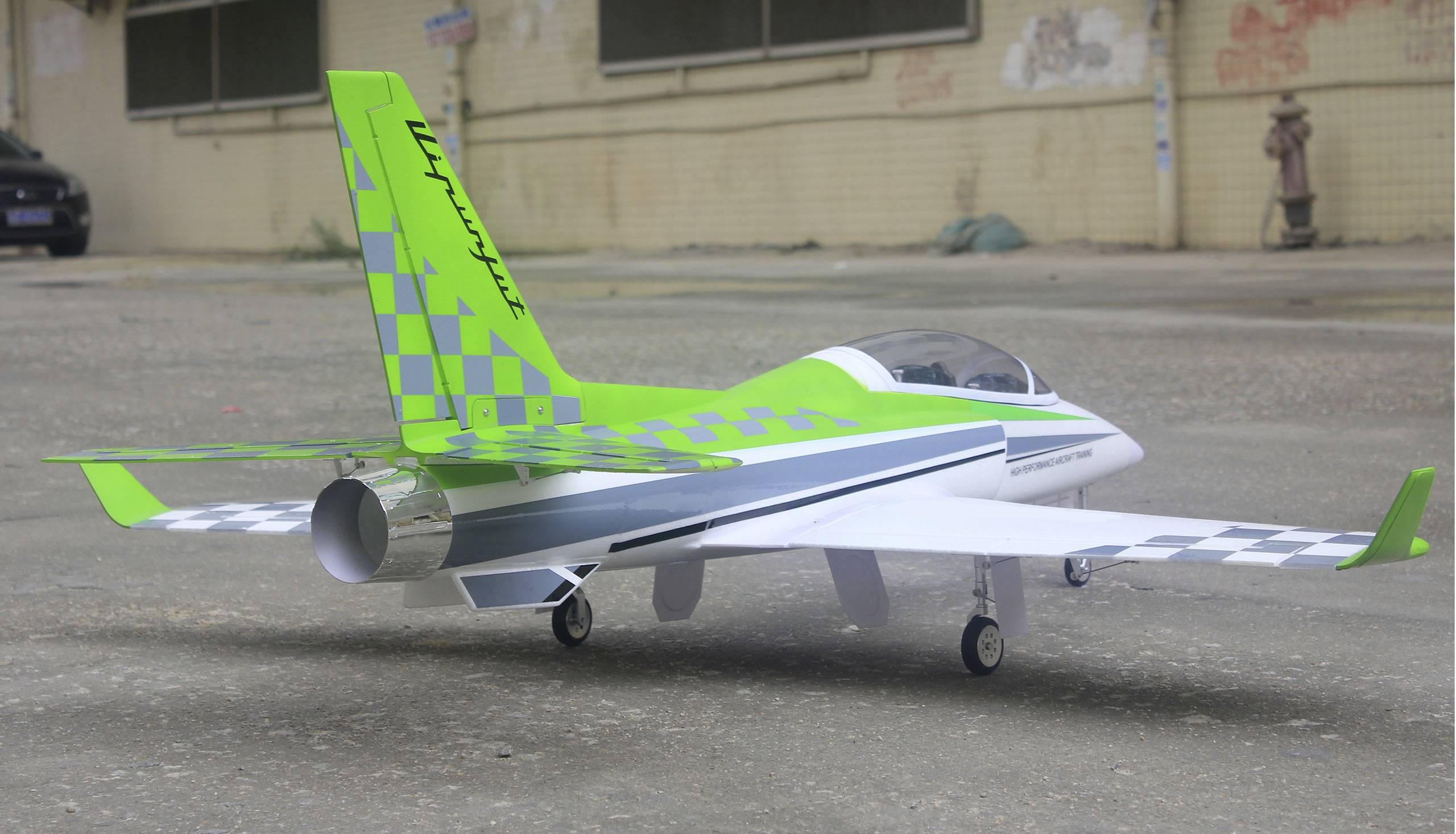 Ein Modellflugzeug in leuchtendem Grün und Weiß mit karierten Mustern steht auf einer Betonfläche vor einem Gebäude im Freien.