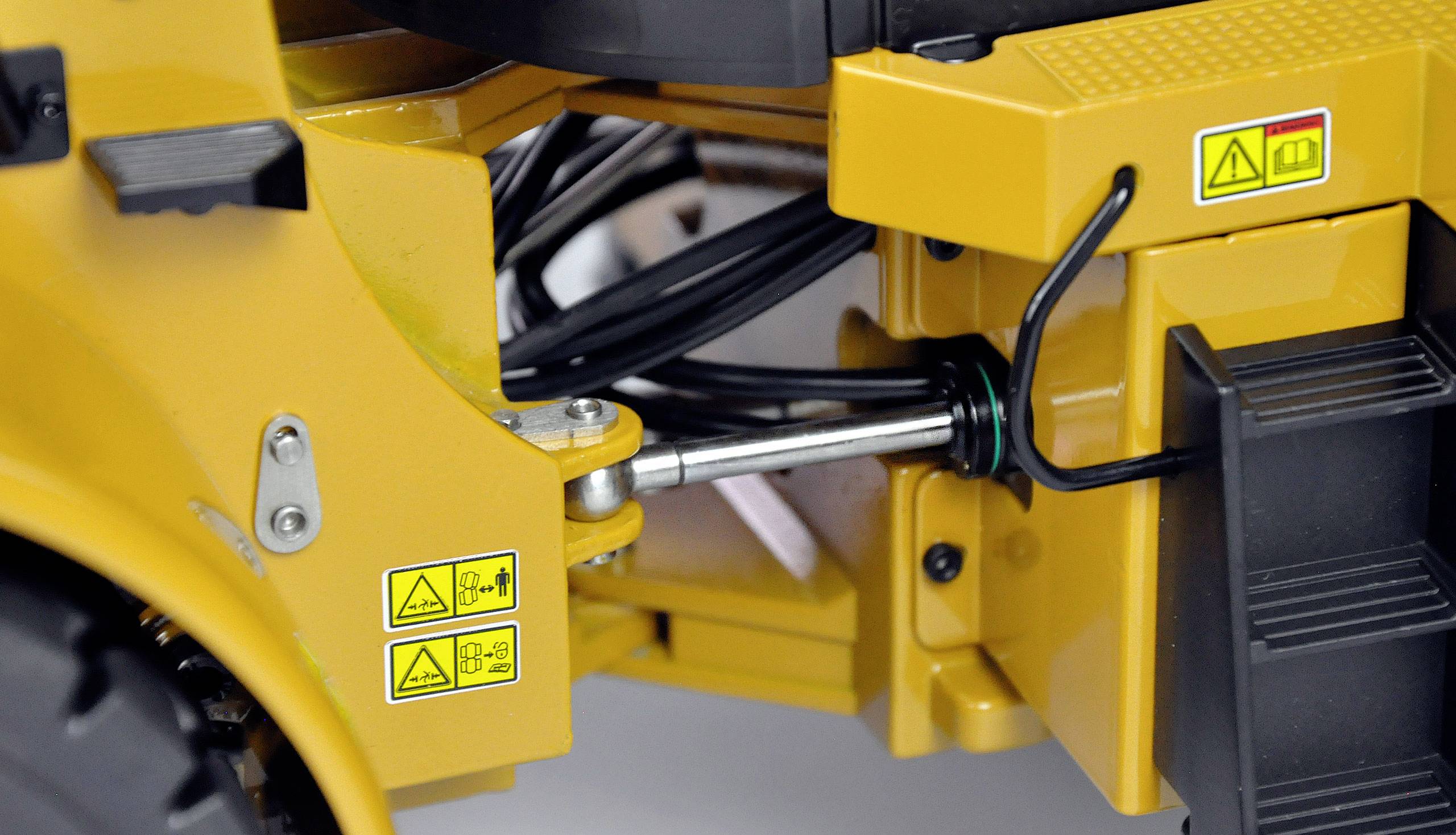 Nahaufnahme eines gelben Industriemaschinenarms mit sichtbaren Hydraulikleitungen und Warnsymbolen.