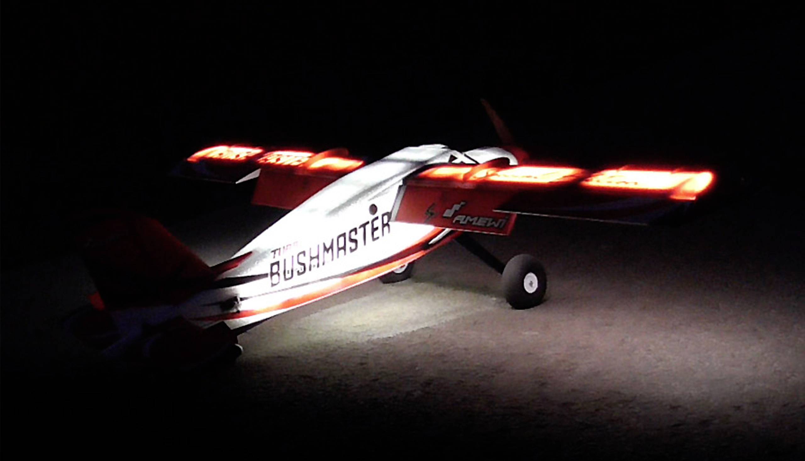 Ein ferngesteuertes Modellflugzeug mit leuchtenden Flügellichtern ist in der Dunkelheit zu sehen. Das Modell trägt die Aufschrift 'Bushmaster'.