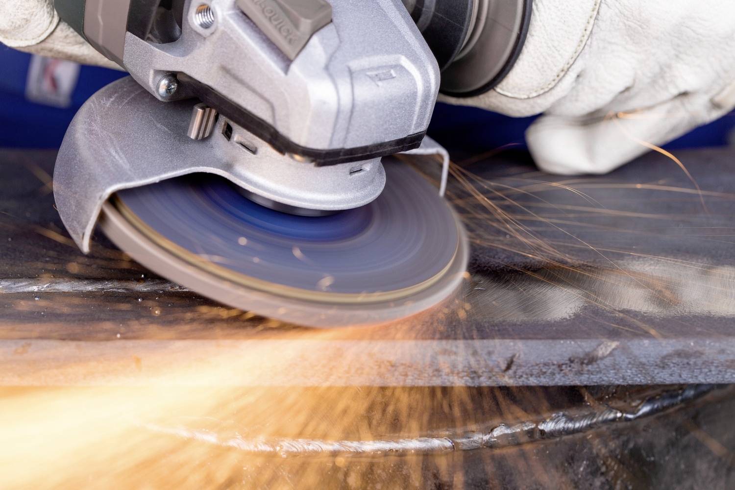 Arbeiter schleift Metallstück mit Winkelschleifer, Funken sprühen. Nahaufnahme zeigt Handschuhe und rotierende Scheibe.