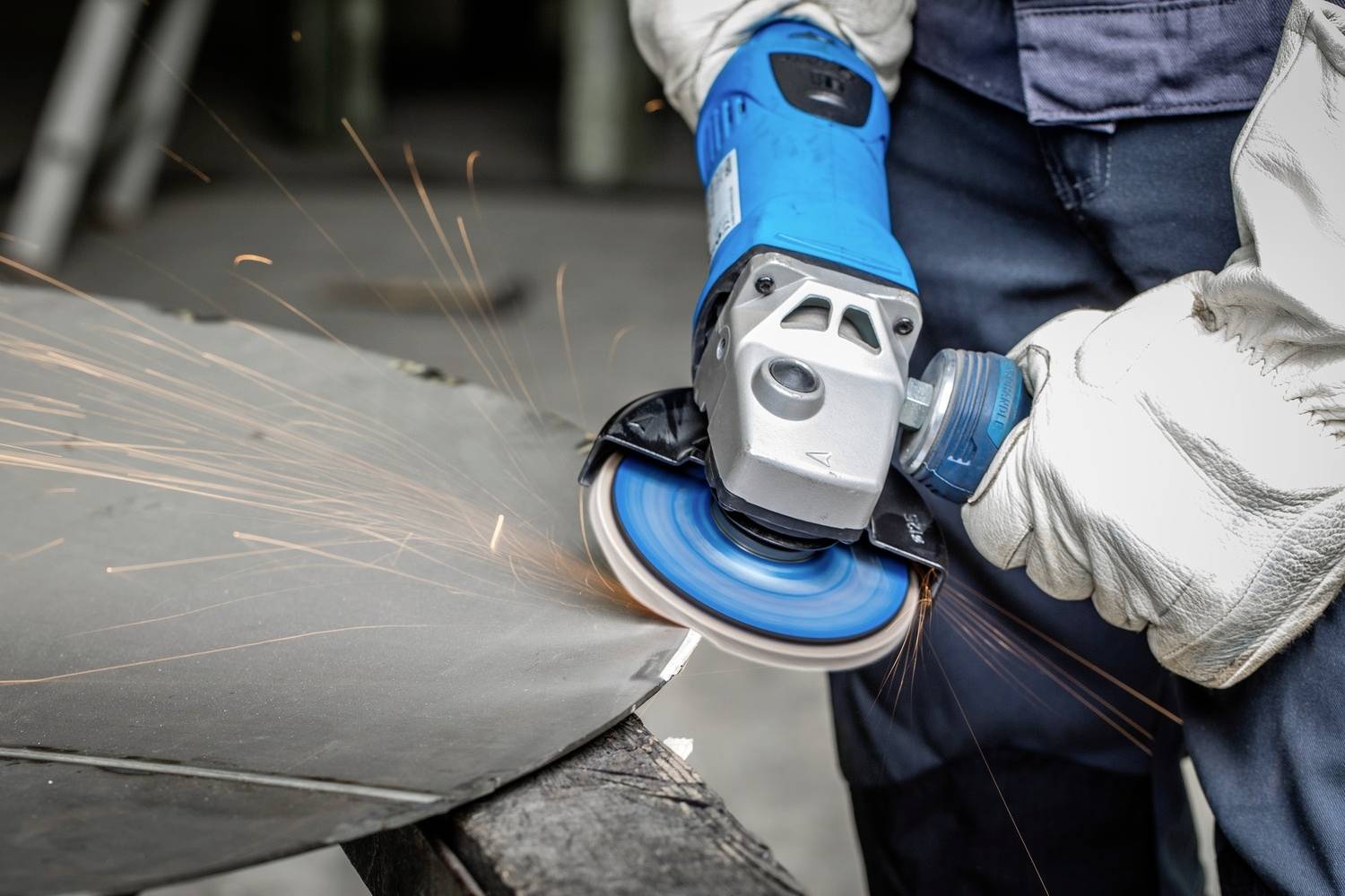 Person schleift ein Metallstück mit einem Winkelschleifer, Funken fliegen. Sie trägt Arbeitshandschuhe für Sicherheit.