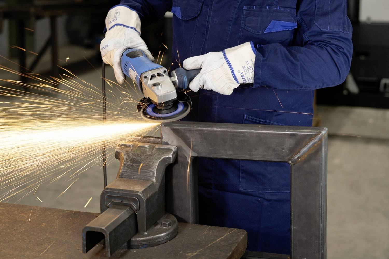 Eine Person in Schutzkleidung schleift Metall mit einem Winkelschleifer, Funken fliegen.